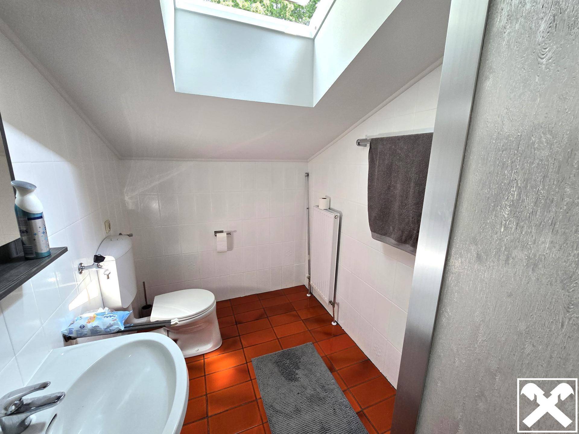 Helles Badezimmer mit Dachfenster, weißen Fliesen und Terrakotta-Boden, ausgestattet mit Toilette und Waschbecken.