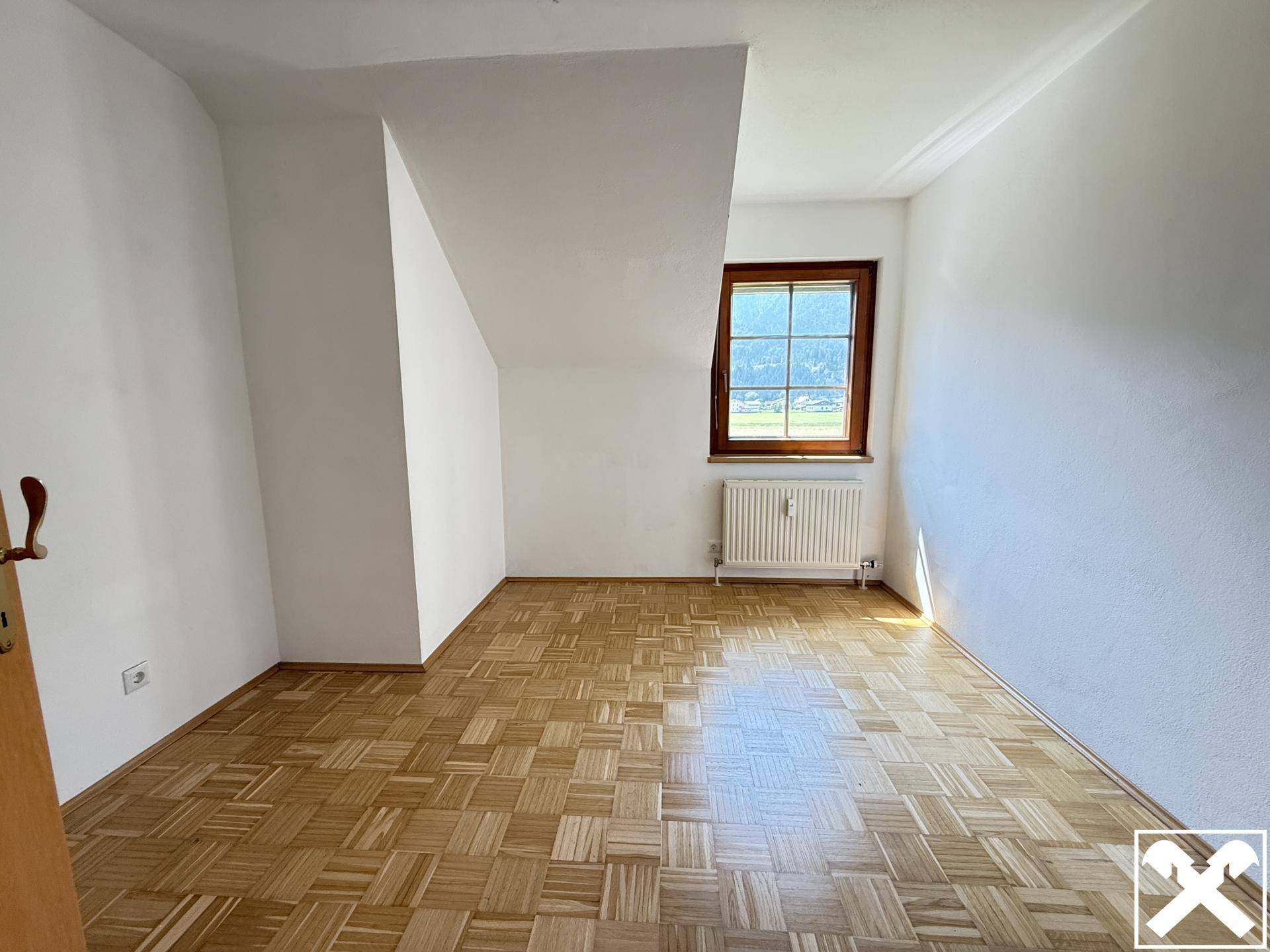 Leeres Zimmer mit Parkettboden und schrägen Wänden, bietet viel natürliches Licht durch das Fenster.