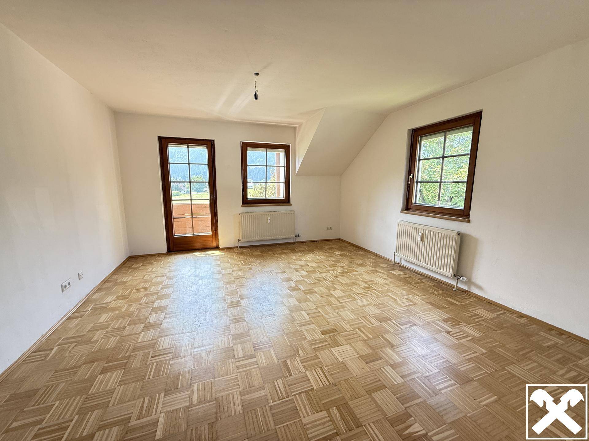 Helles Zimmer mit Parkettboden, Fenster und Zugang zu einem Balkon, unter einem schrägen Dach.
