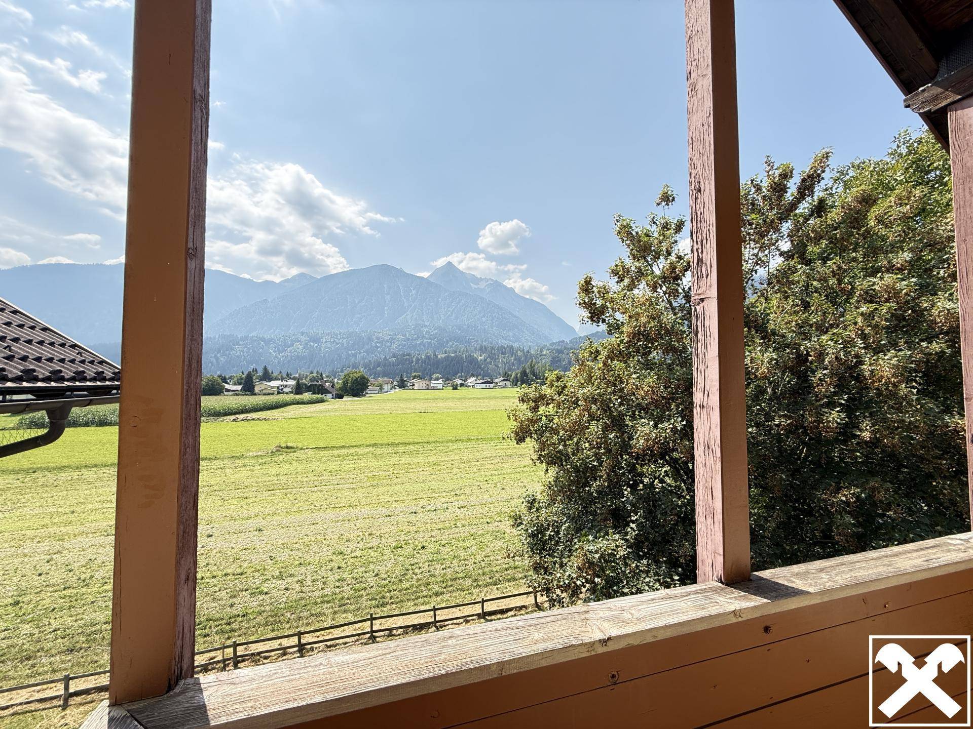Blick von einem Balkon über grüne Felder und majestätische Berge unter blauem Himmel.