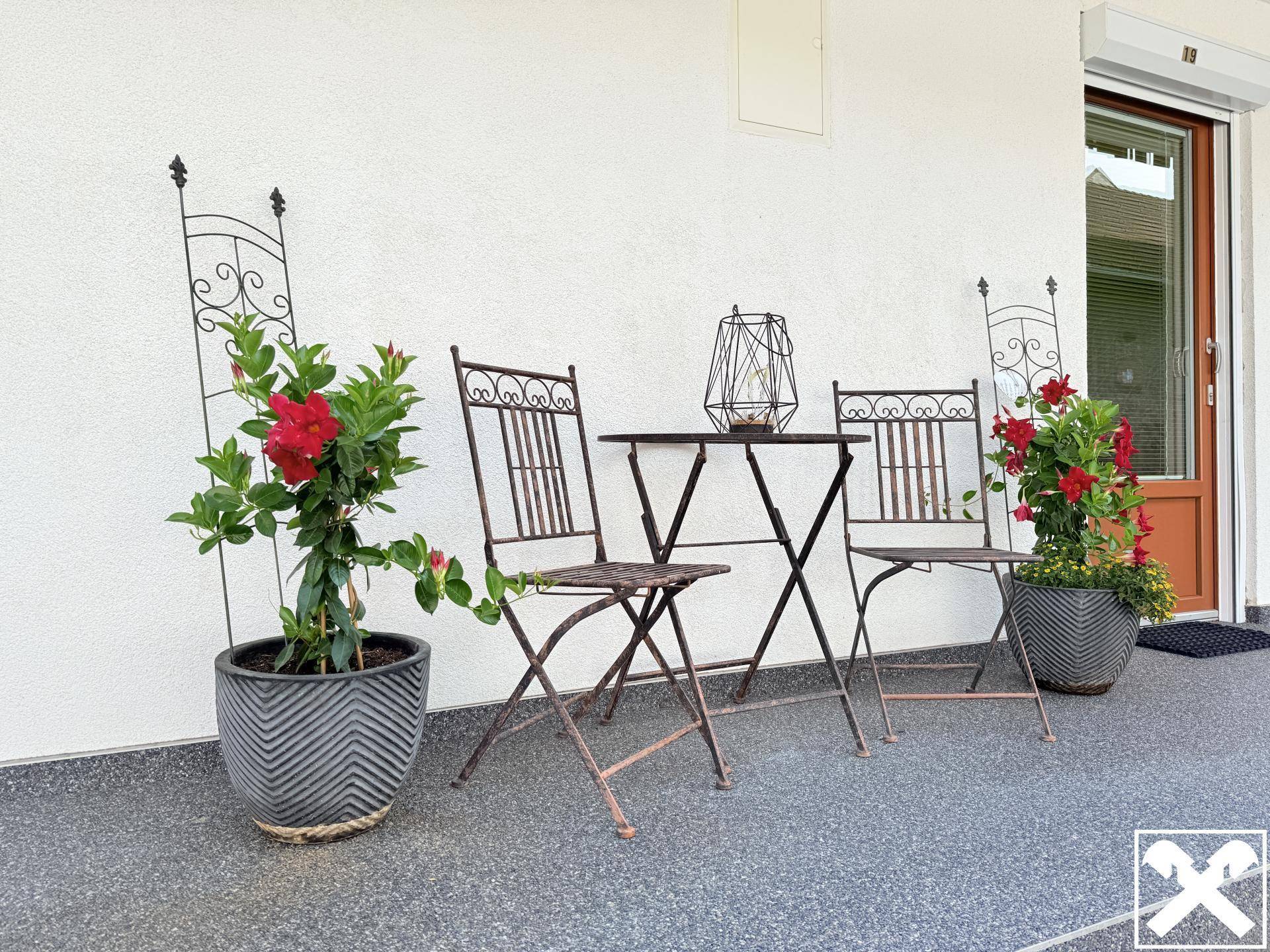 Kleine Terrasse mit schmiedeeisernen Möbeln und blühenden Pflanzen in Töpfen.