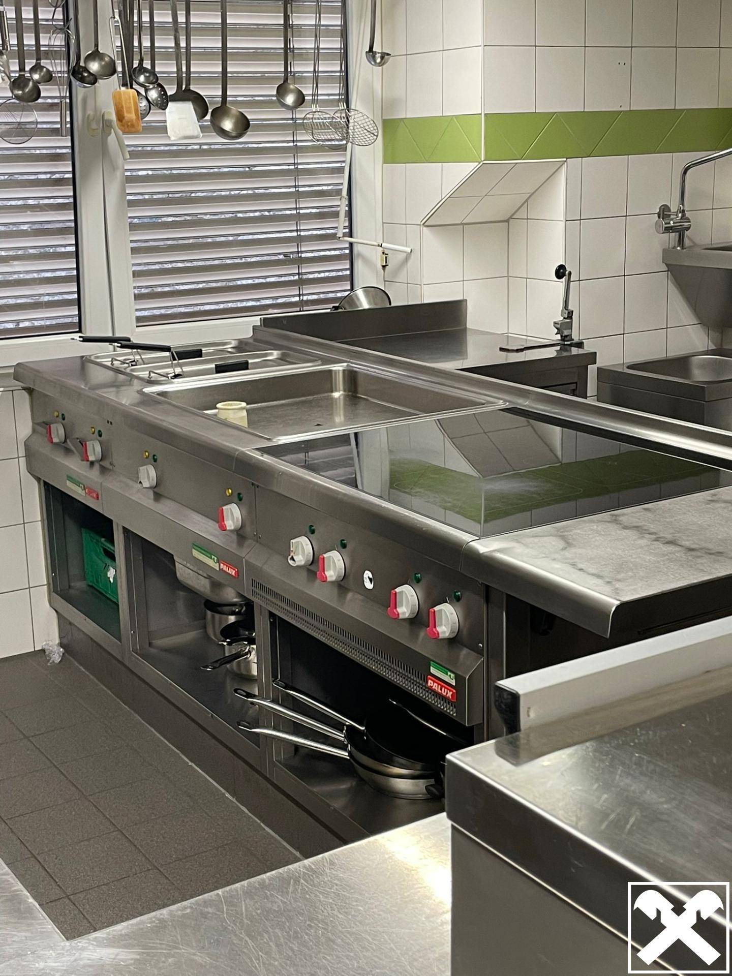 Professionelle Großküche mit Edelstahlgeräten, Induktionskochfeldern und Hängeregalen für Utensilien.