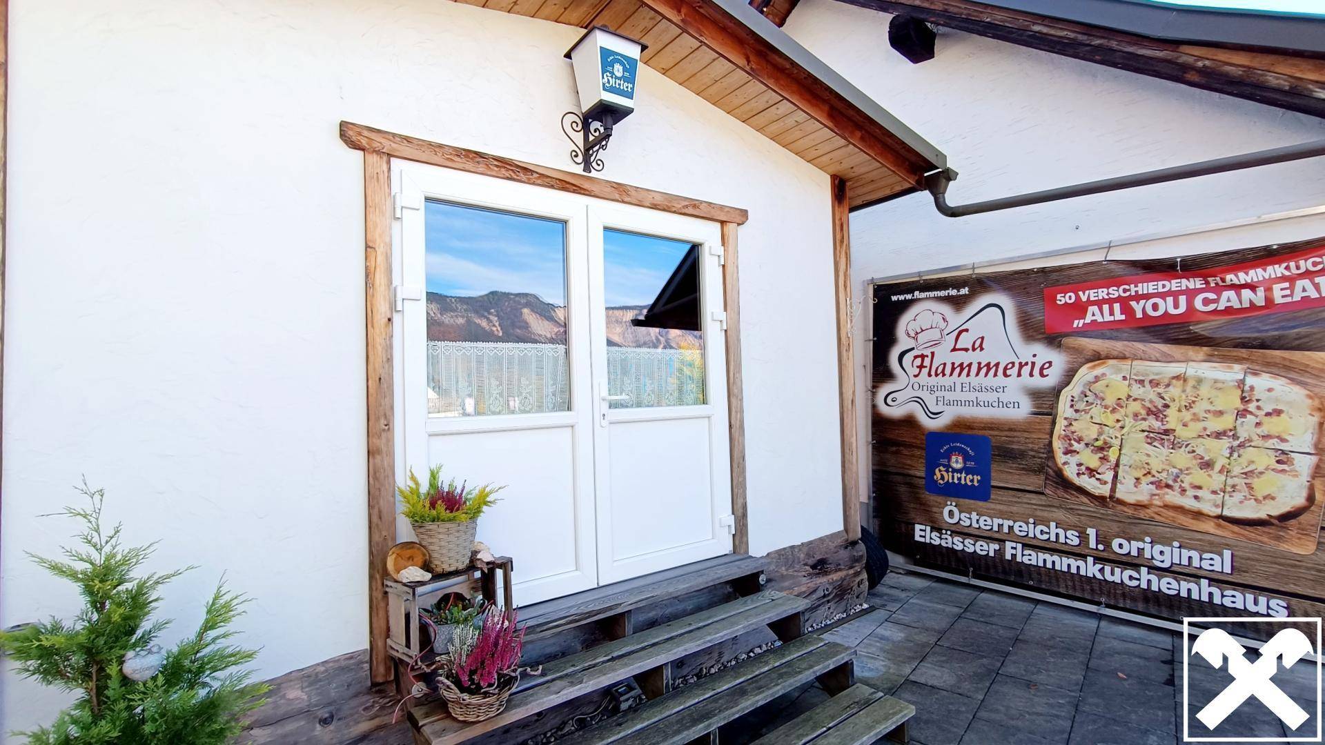 Eingangsbereich des Gebäudes mit einer weißen Doppeltür, rustikalen Holzrahmen und einer Werbetafel für das Restaurant.