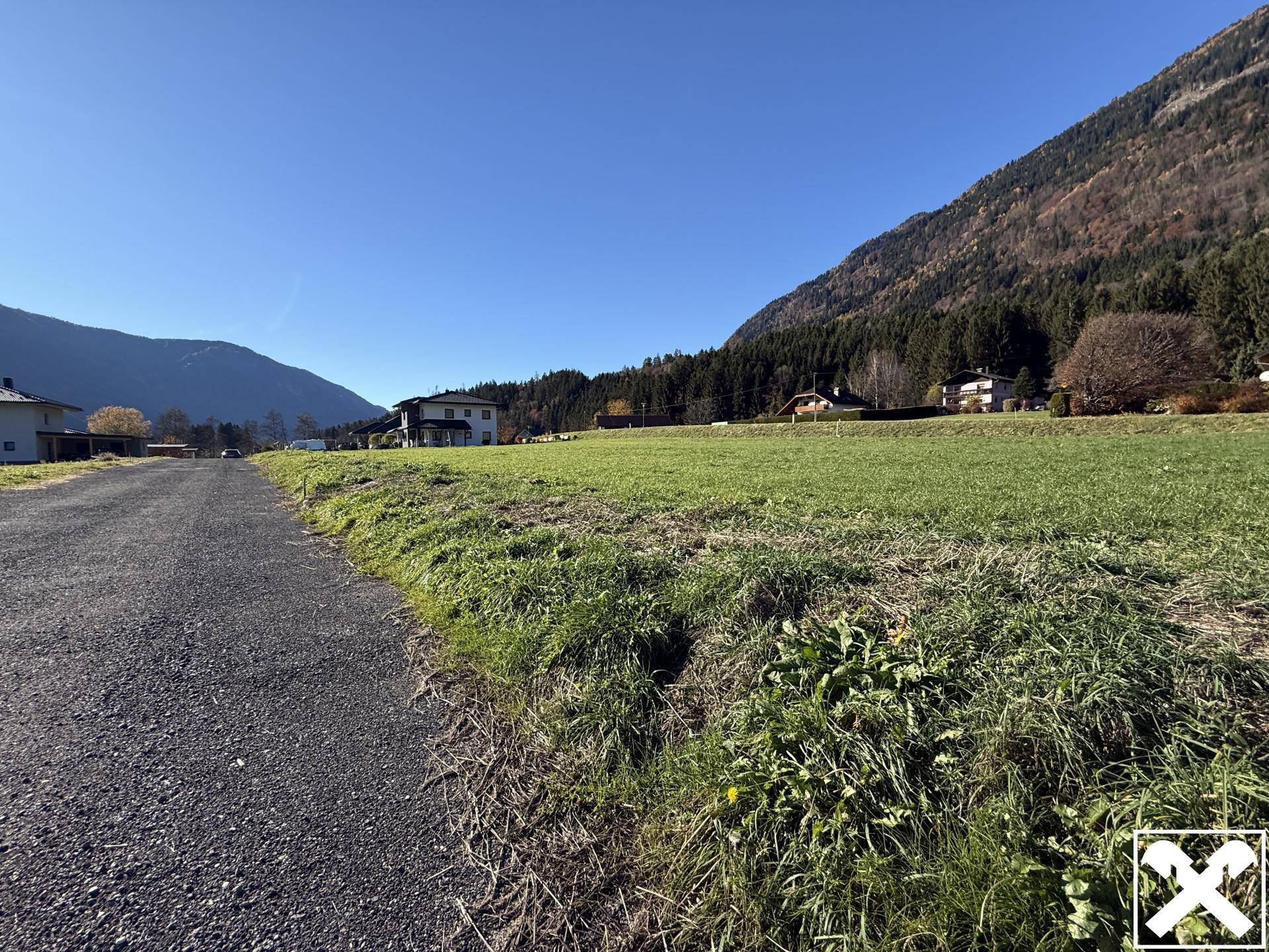 Schotterweg neben einer Wiese mit Wohnhäusern und bewaldeten Bergen im Hintergrund.