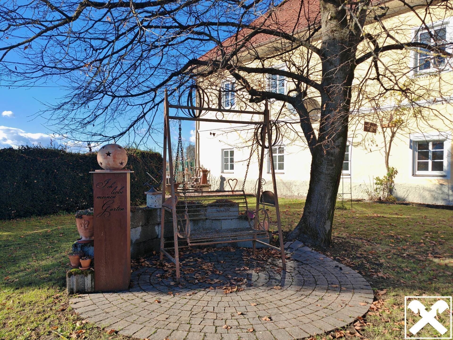 Gartenbereich mit einer Schaukel, gepflastertem Sitzplatz und altem Baumbestand.