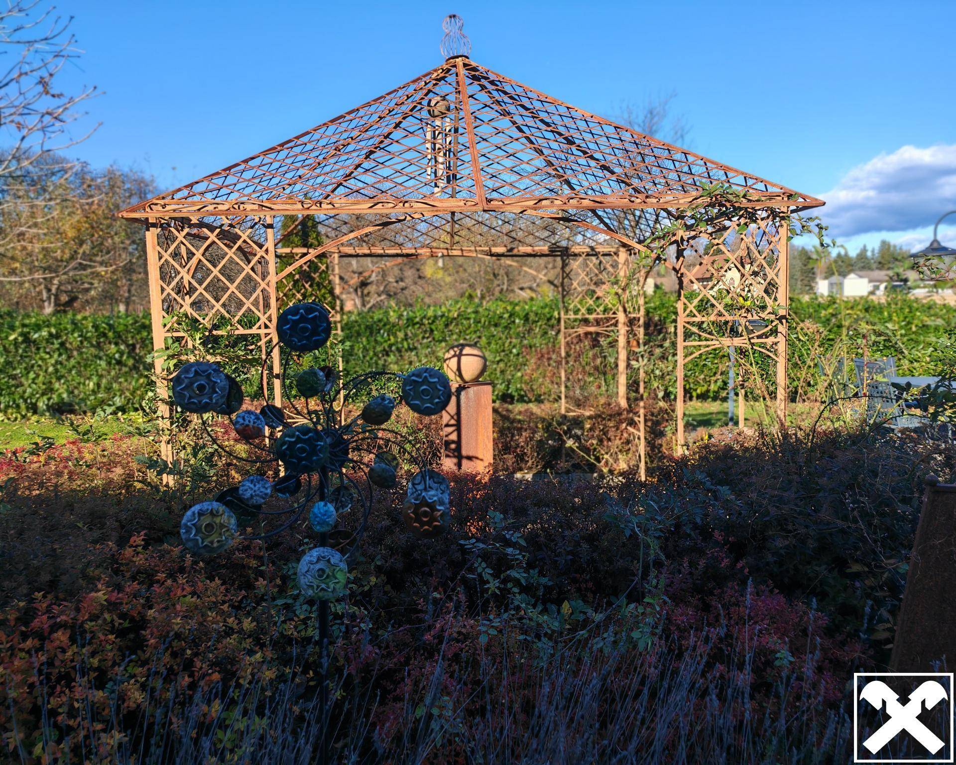 Dekorativer Gartenpavillon aus Metall, umgeben von Sträuchern und Blumenbeeten.