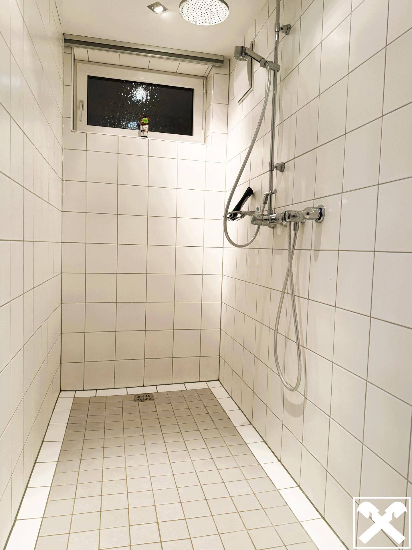 Helle Duschkabine mit weißen Fliesen, Regendusche und einem Fenster für natürliche Belüftung.