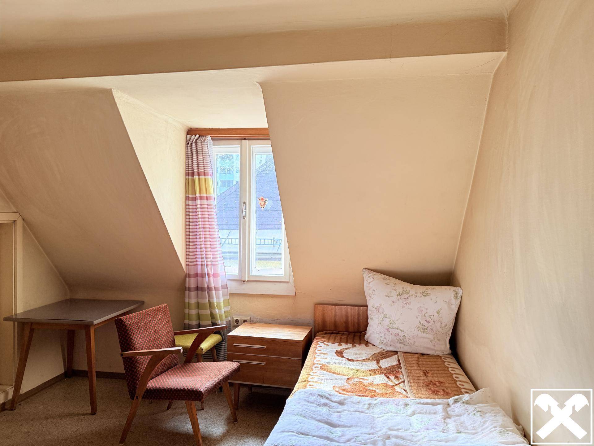 Dachschlafzimmer mit schrägen Decken, Einzelbett und einem Fenster mit bunten Vorhängen, das viel Licht spendet.