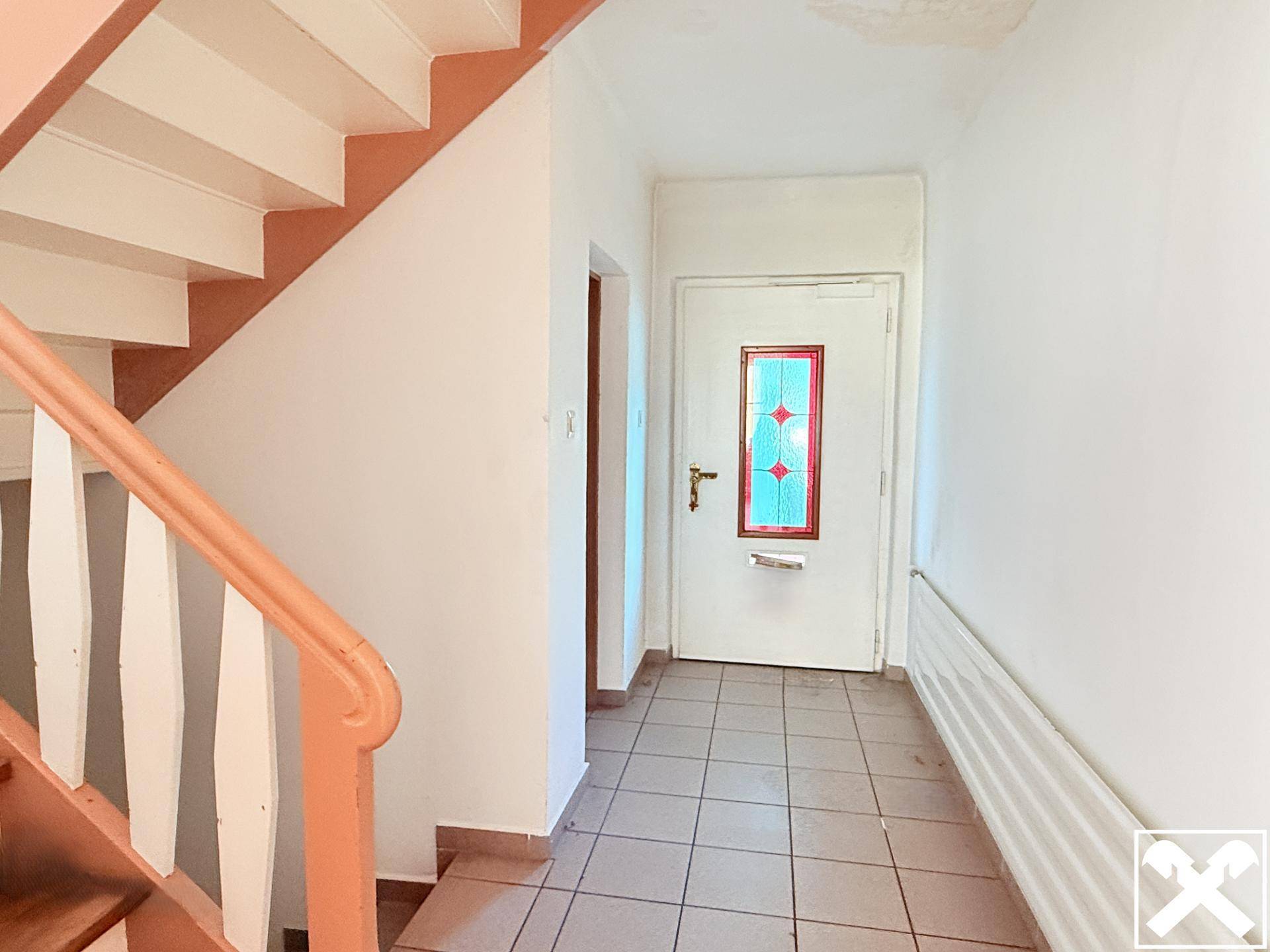 Eingangsbereich mit gefliestem Boden, weißer Tür mit Buntglas und einer Holztreppe, die nach oben führt.