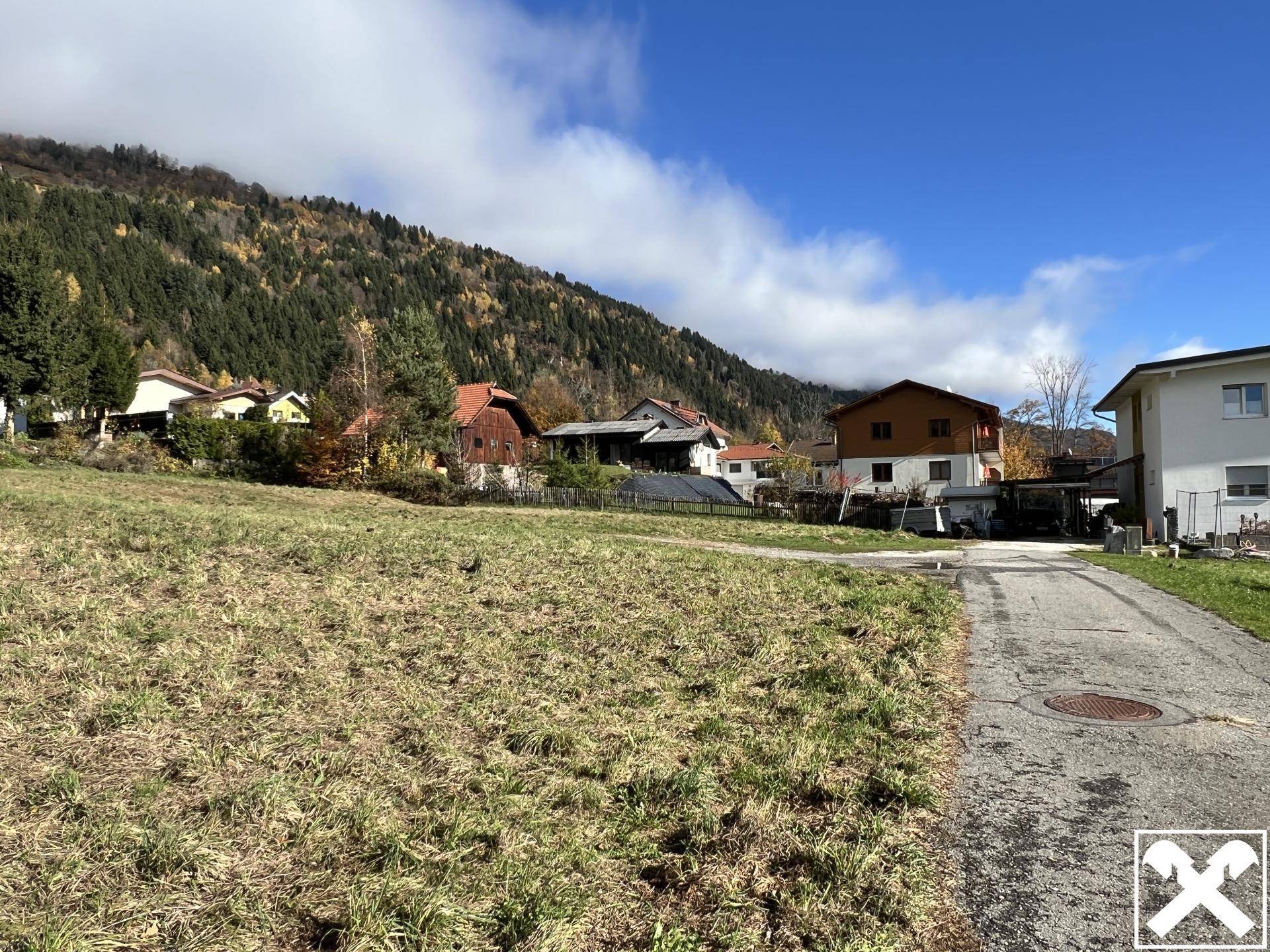 Offenes Grundstück mit Zufahrtsweg und Blick auf die umliegenden Häuser und Berge.