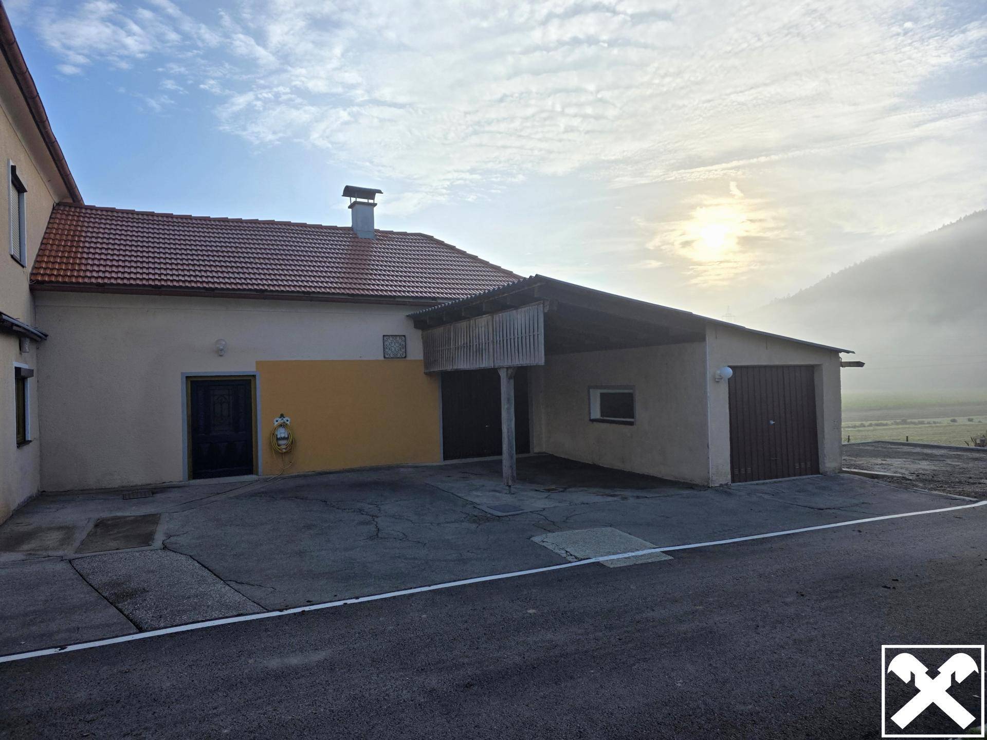 Rückseite des Gebäudes mit Garagen und einem Carport, umgeben von ländlicher Landschaft.