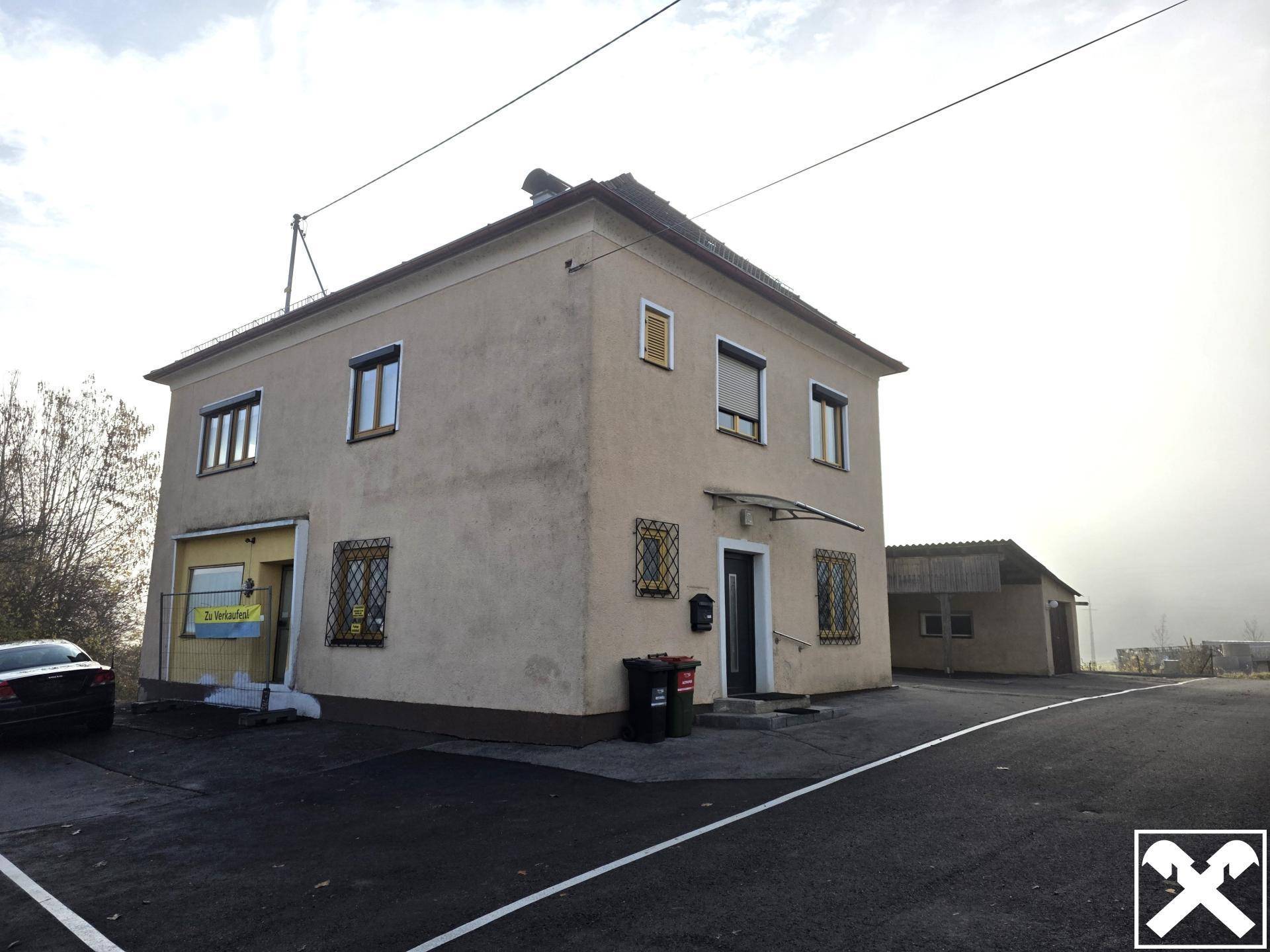 Vorderansicht eines älteren Gebäudes mit beiger Fassade, Parkplätzen und einem Nebengebäude im Hintergrund.