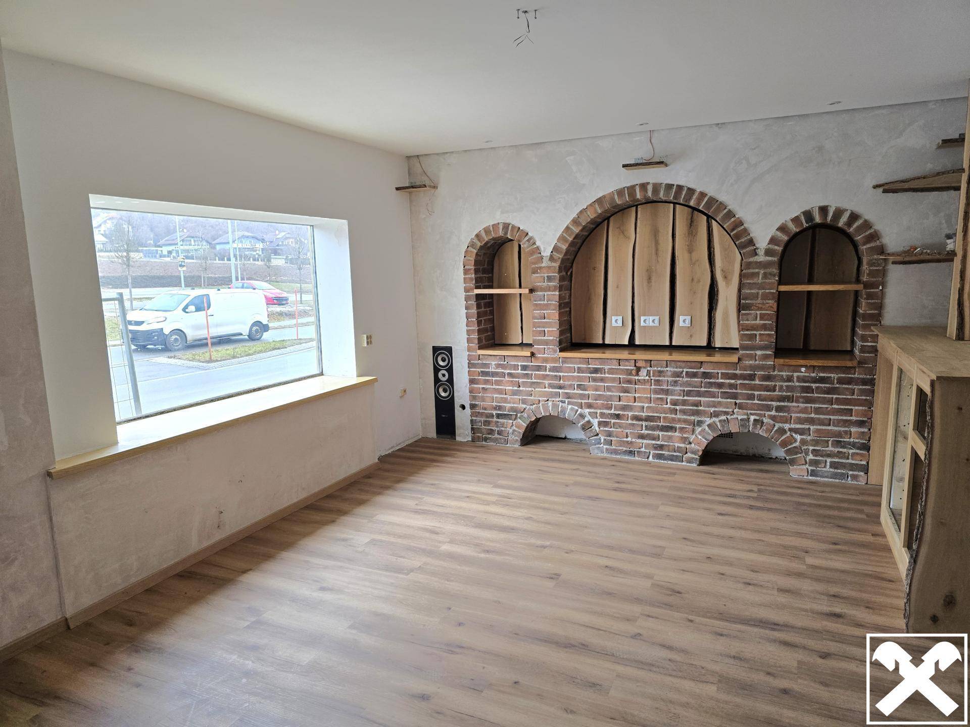 Wohnzimmer mit großem Fenster, Holzboden und dekorativen Ziegelnischen, die Renovierungsbedarf zeigen.