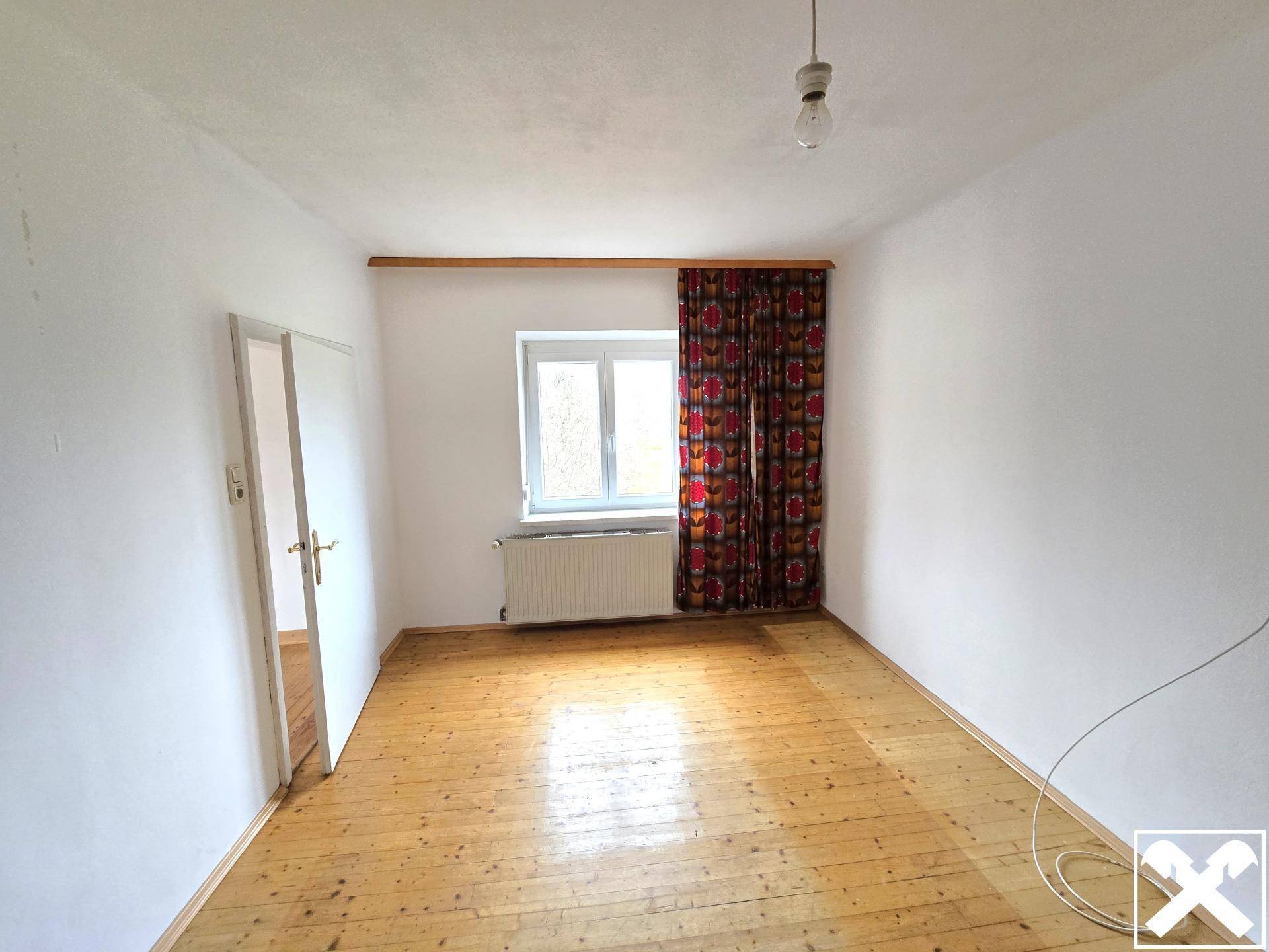 Schlafzimmer mit Holzdielenboden und einem Fenster mit Vorhang, das Renovierungsbedarf zeigt.