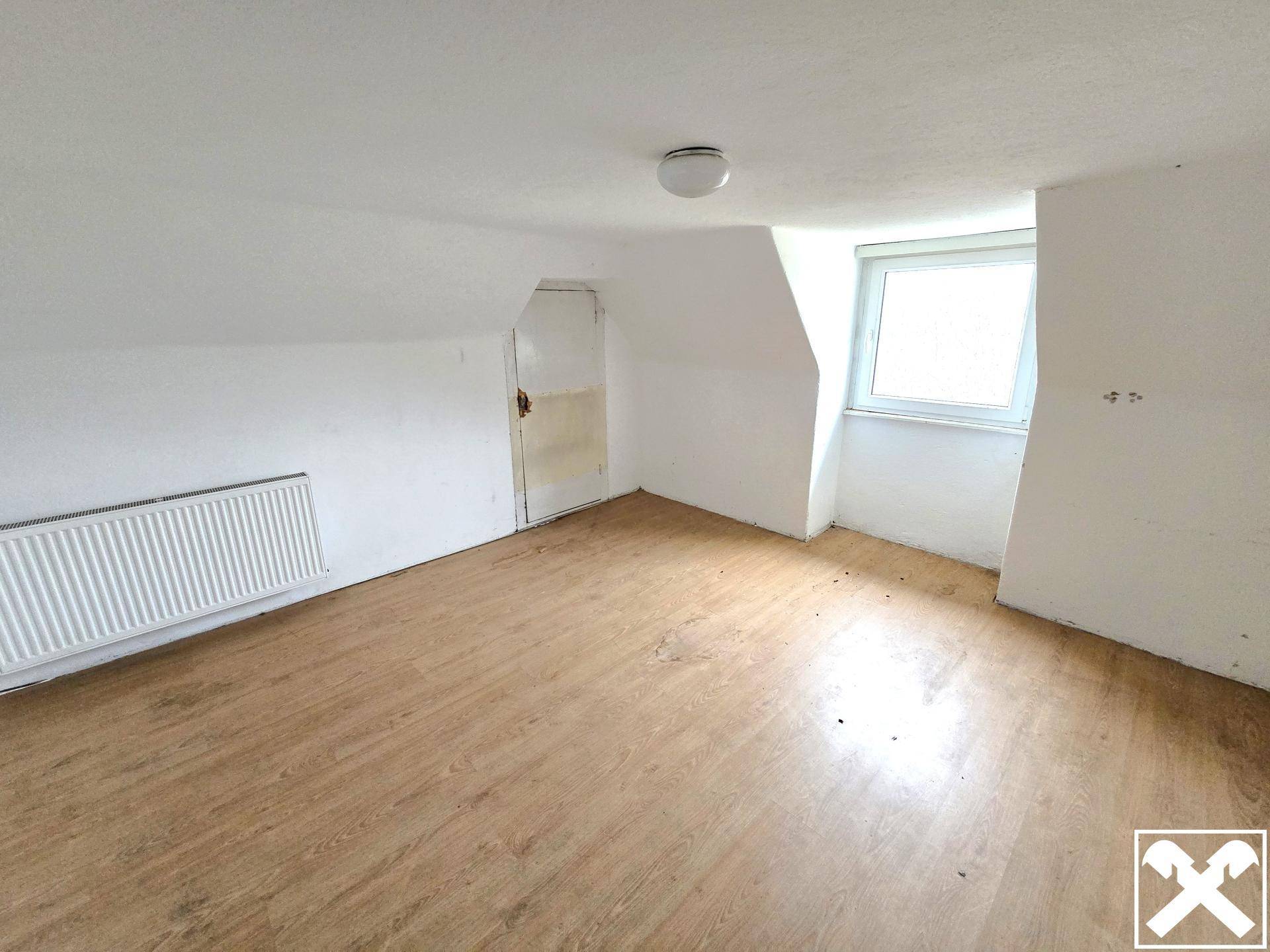 Dachzimmer mit schräger Decke, Laminatboden und einem Fenster, das Renovierungsbedarf zeigt.