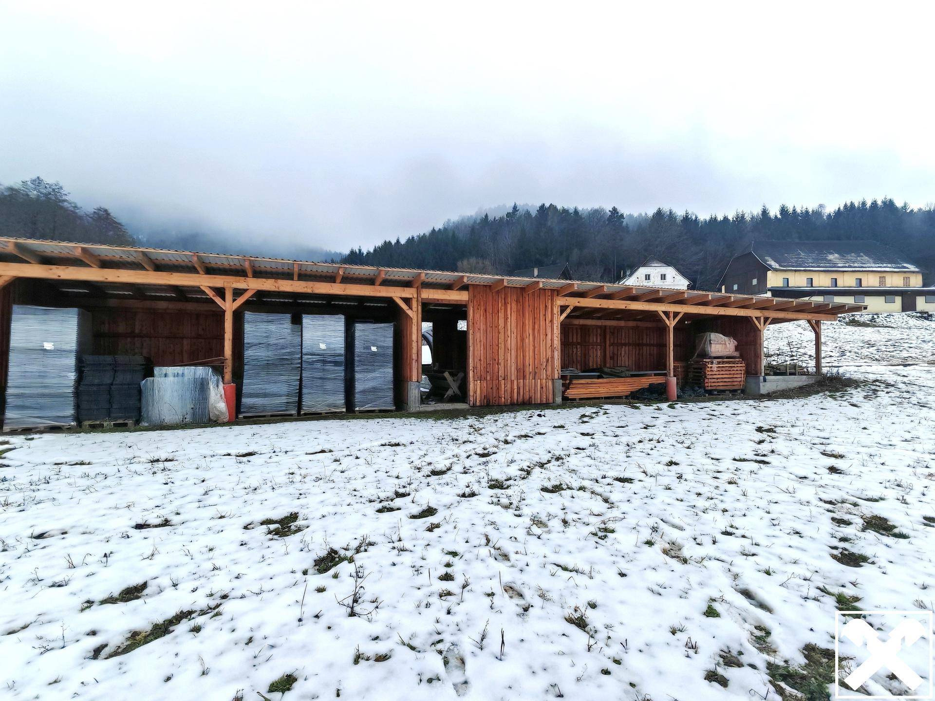 Eine lange hölzerne Überdachung mit Lagerbereichen und Schnee, im Hintergrund ein Wald und Gebäude.