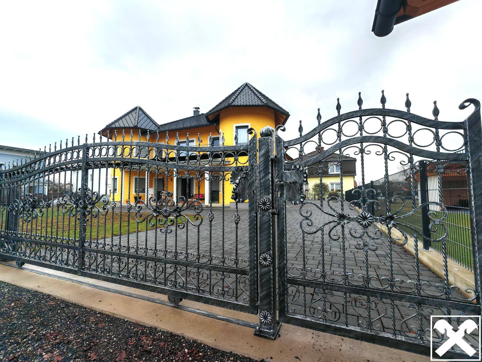 Schmiedeeisernes Tor vor dem gelben Haus mit gepflasterter Auffahrt und Garten.