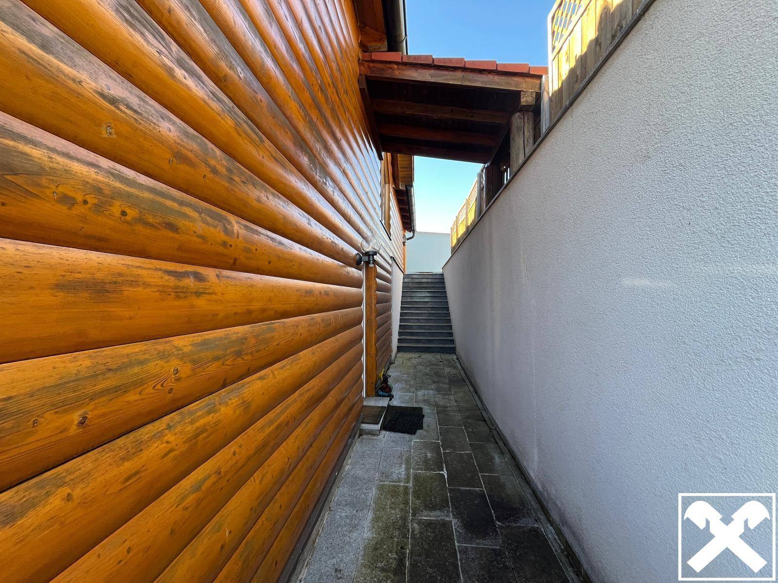 Schmaler gepflasterter Weg zwischen der Holzwand des Hauses und einer weißen Mauer.