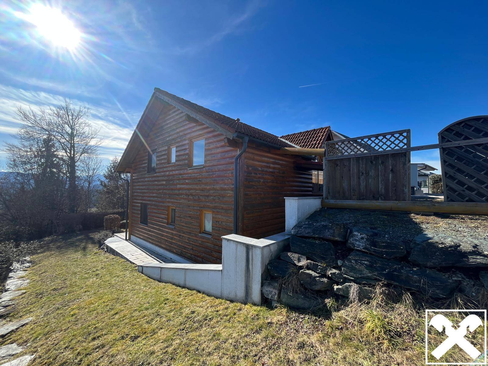 Seitenansicht des Holzhauses mit Felsen und Grünflächen unter strahlendem Sonnenschein.
