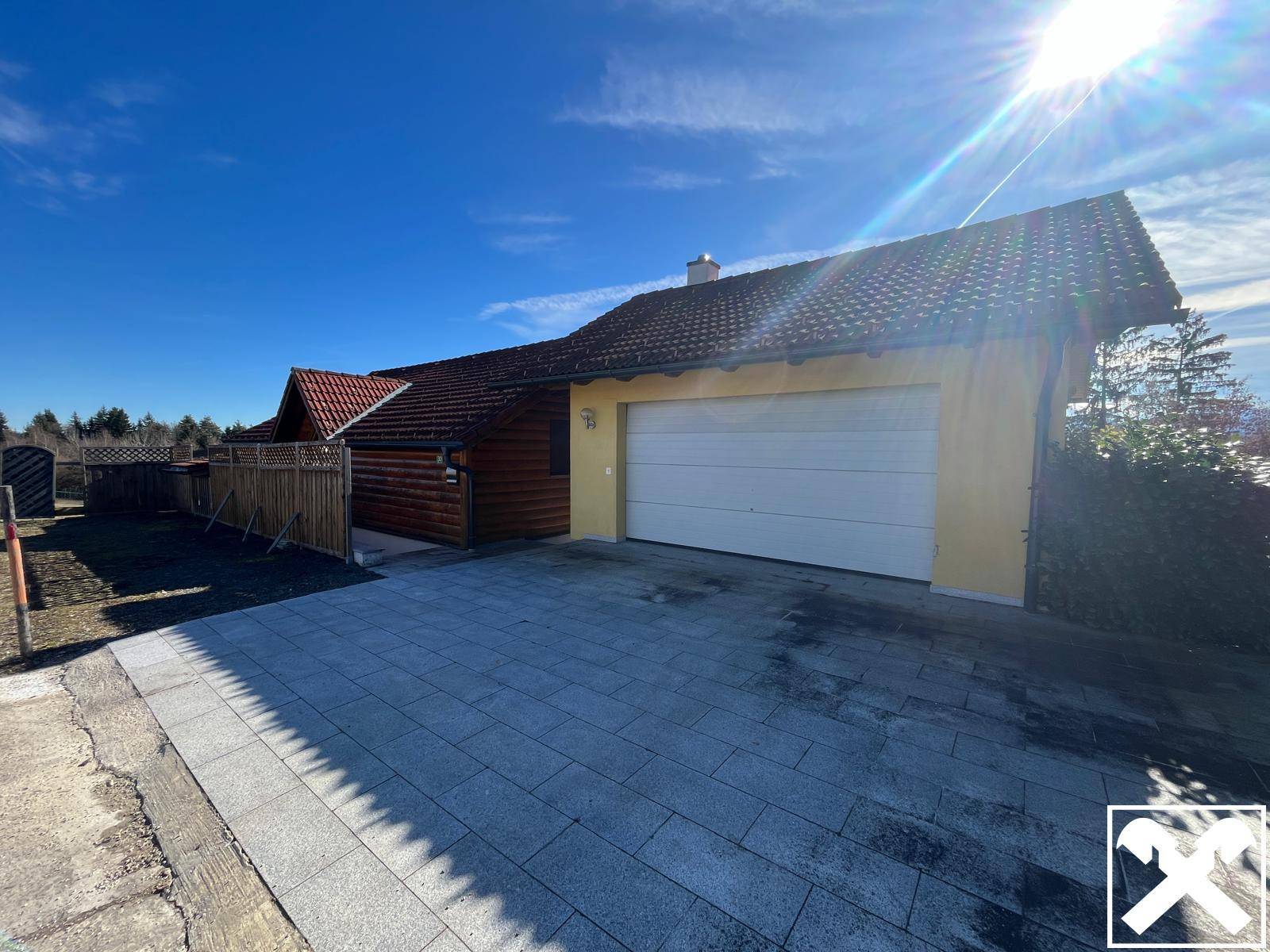 Garage mit weißem Tor und gepflasterter Einfahrt neben dem Holzhaus an einem sonnigen Tag.