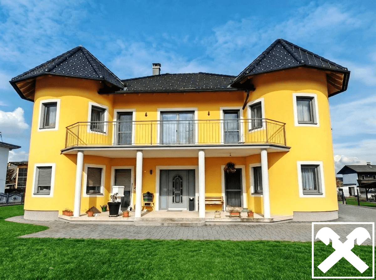 Vorderansicht des gelben Hauses mit Balkon, Säulen und gepflegtem Vorgarten bei Sonnenschein.