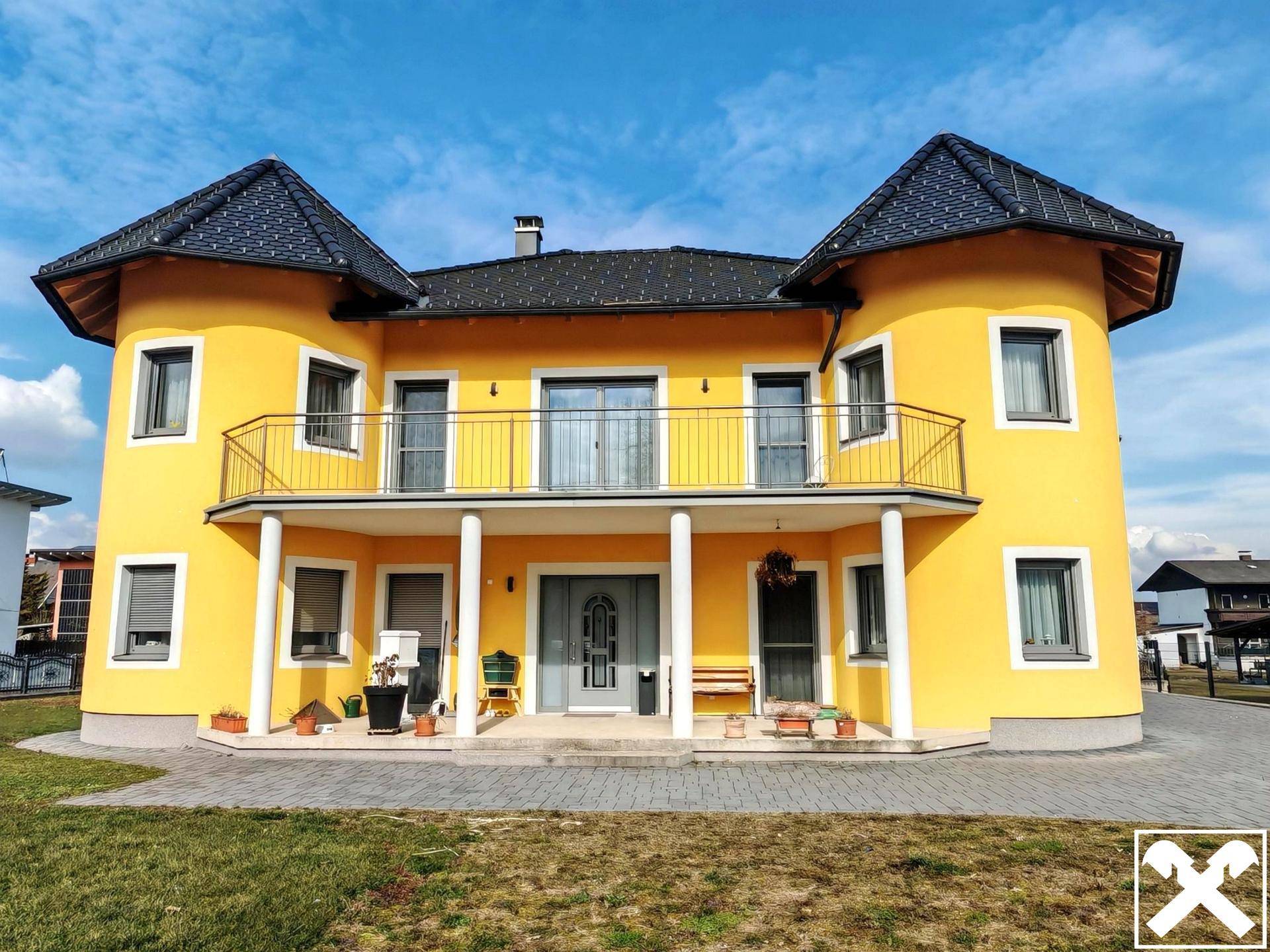 Vorderansicht des gelben Hauses mit Balkon, Säulen und gepflegtem Vorgarten bei Sonnenschein.