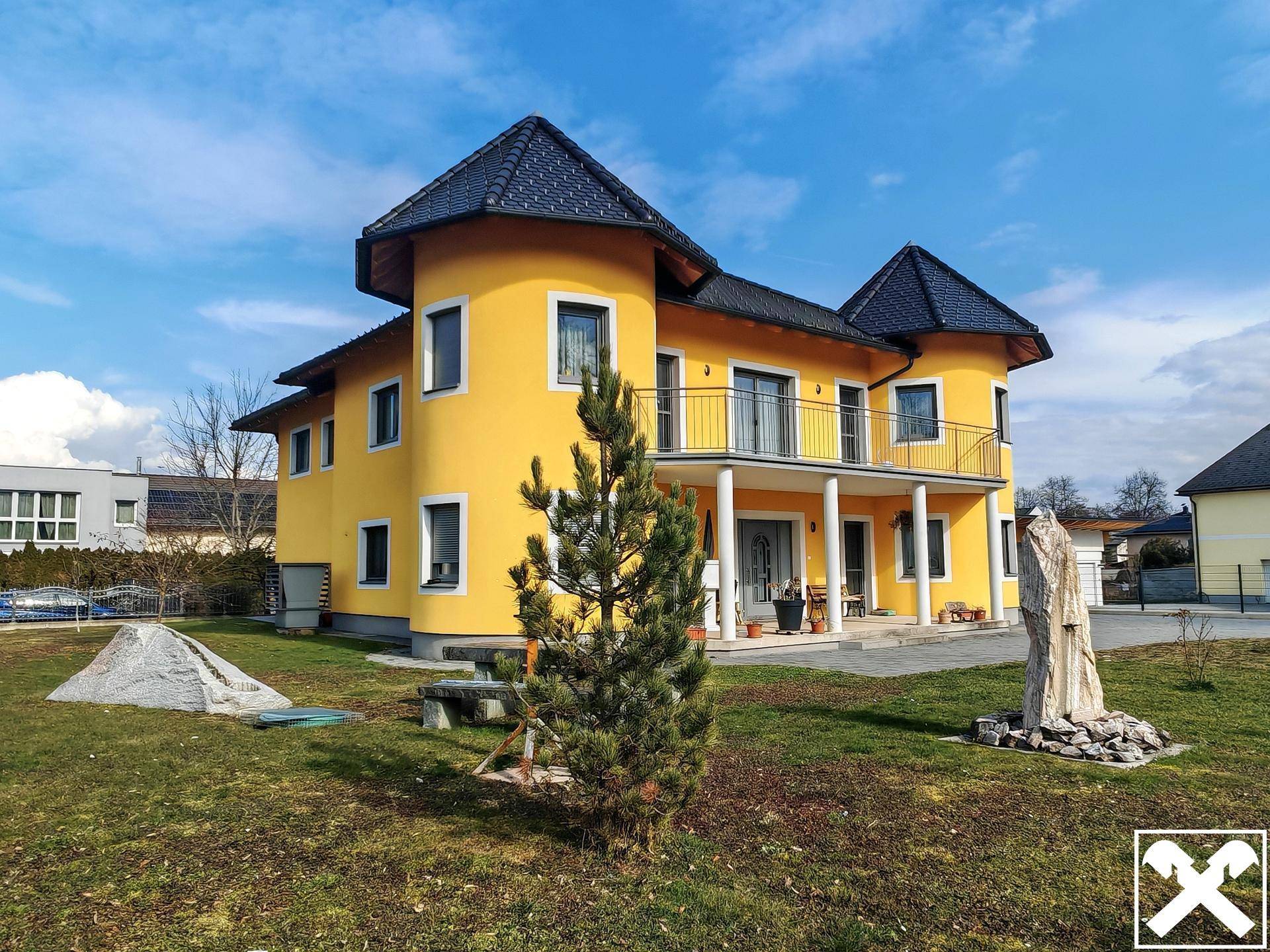 Gelbes Haus mit Garten, gepflasterter Auffahrt und dekorativen Elementen unter blauem Himmel.