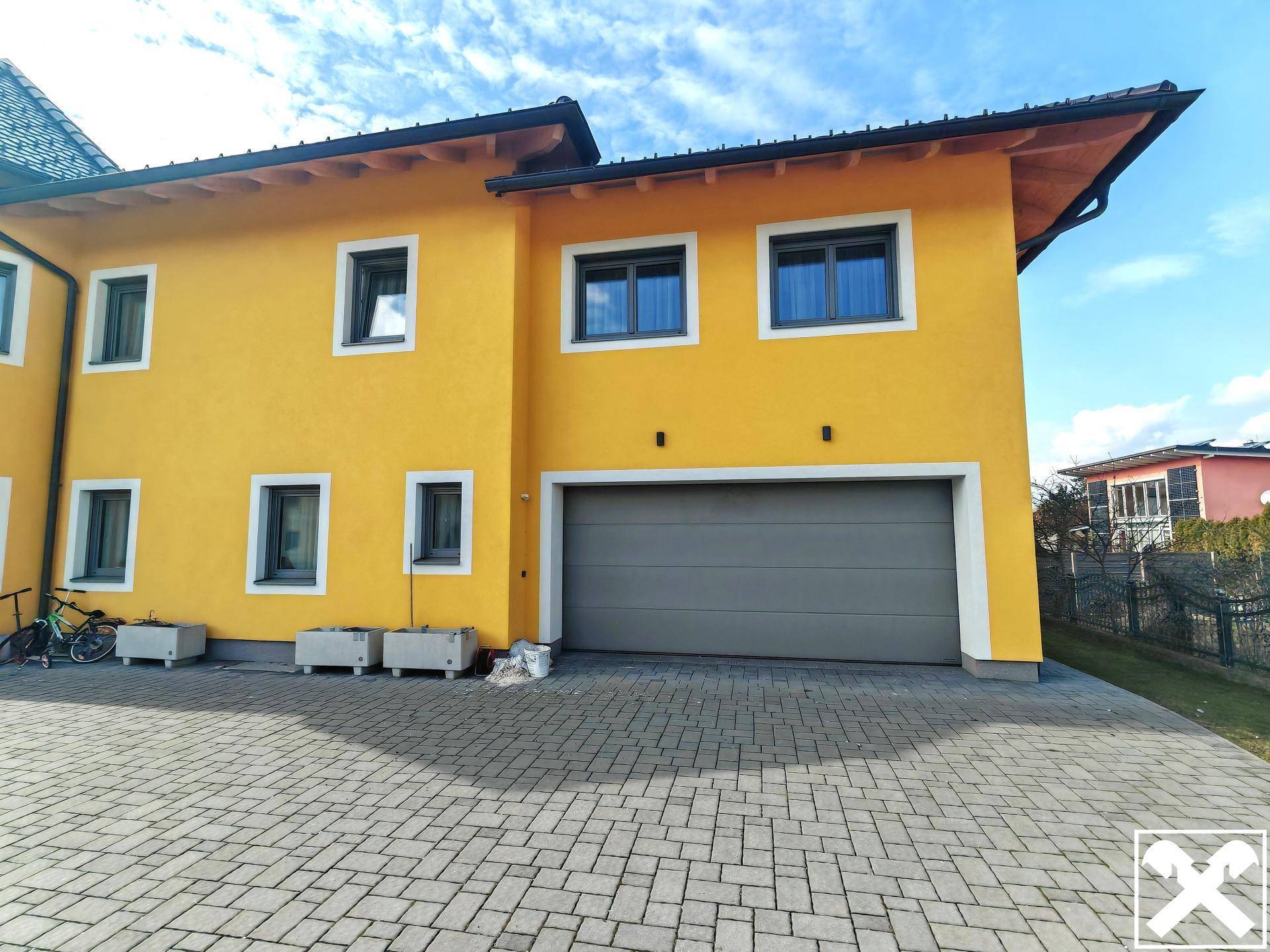 Seitenansicht des gelben Hauses mit Garage und gepflasterter Auffahrt bei Sonnenschein.