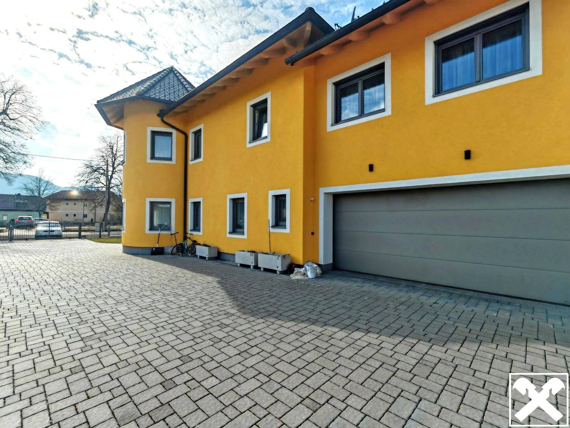 Seitenansicht des gelben Hauses mit Garage und gepflasterter Auffahrt unter blauem Himmel.