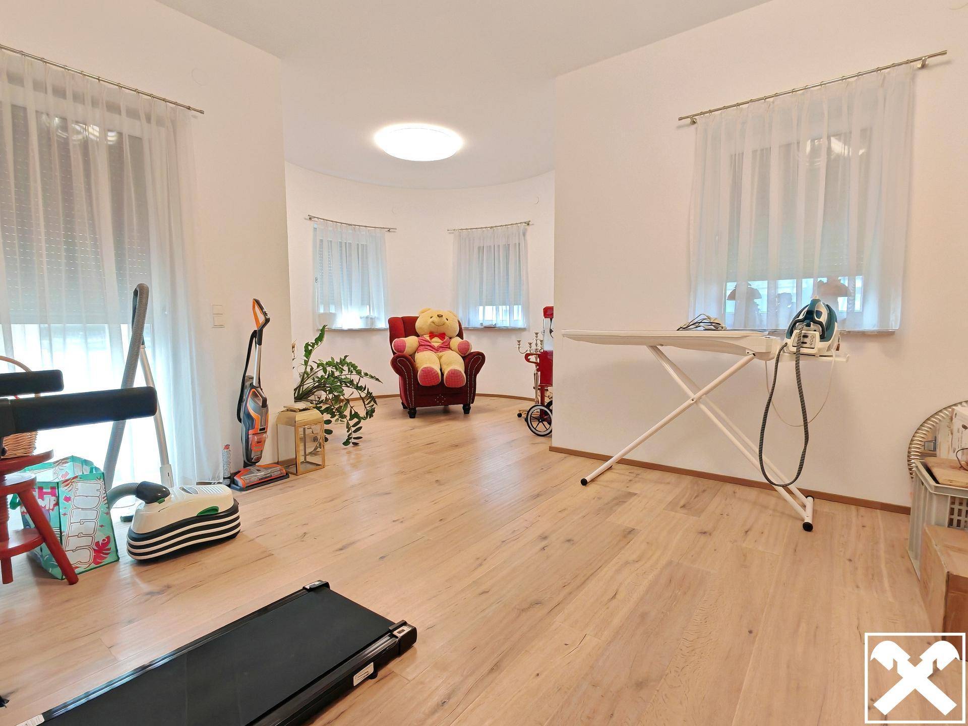 Heller Mehrzweckraum mit Holzboden, mehreren Fenstern und verschiedenen Einrichtungsgegenständen.