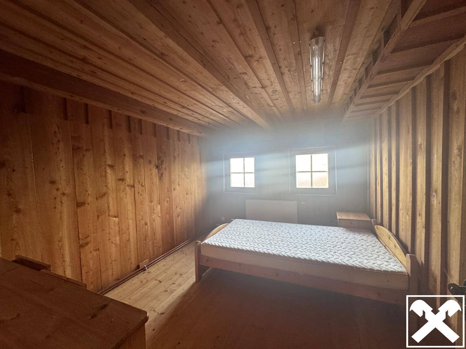 Schlafzimmer mit Holzvertäfelung, Holzboden und einem Bett unter zwei Fenstern.