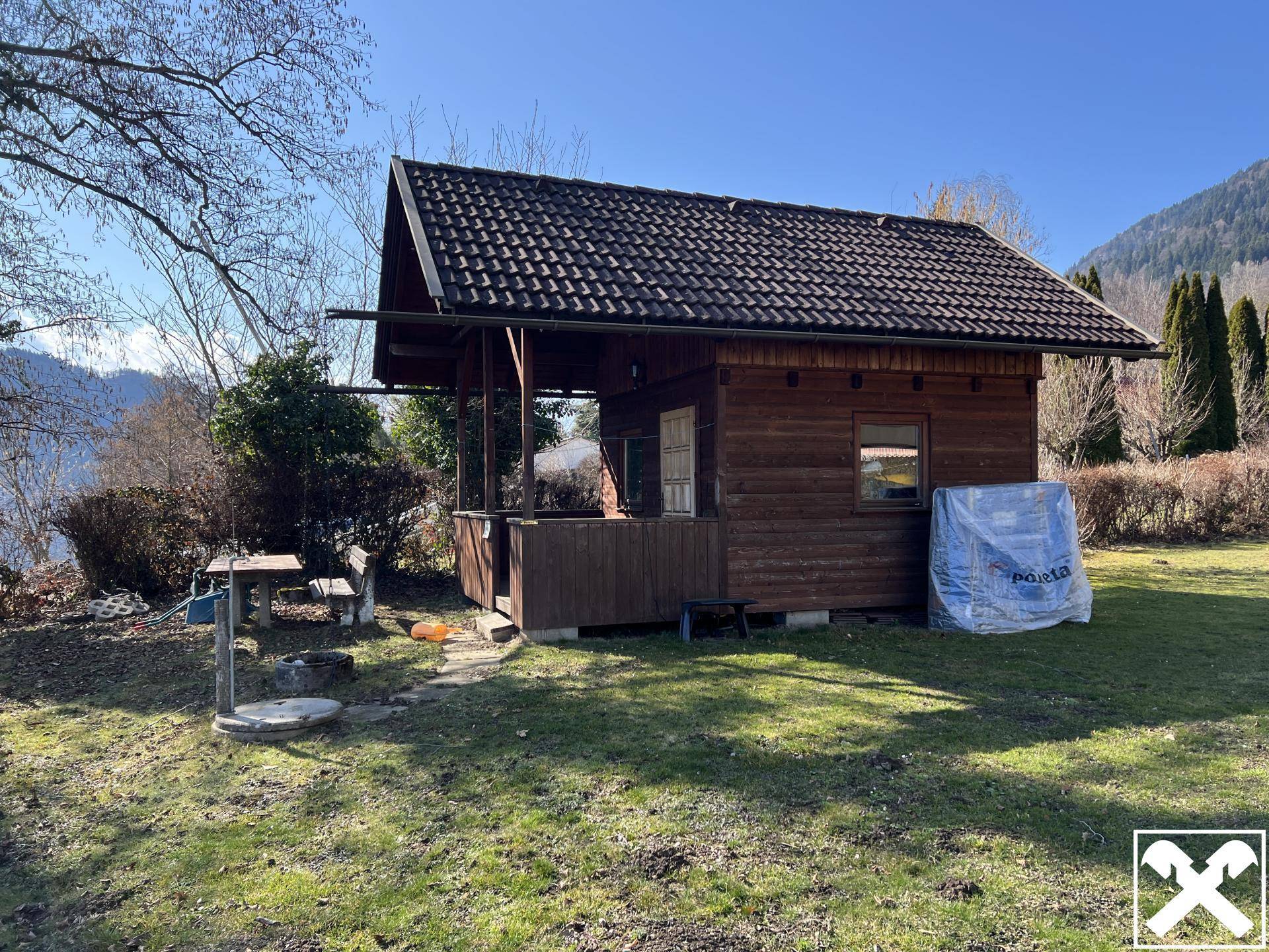 Rustikales Holzhaus mit überdachter Veranda und Feuerstelle im sonnigen Garten.