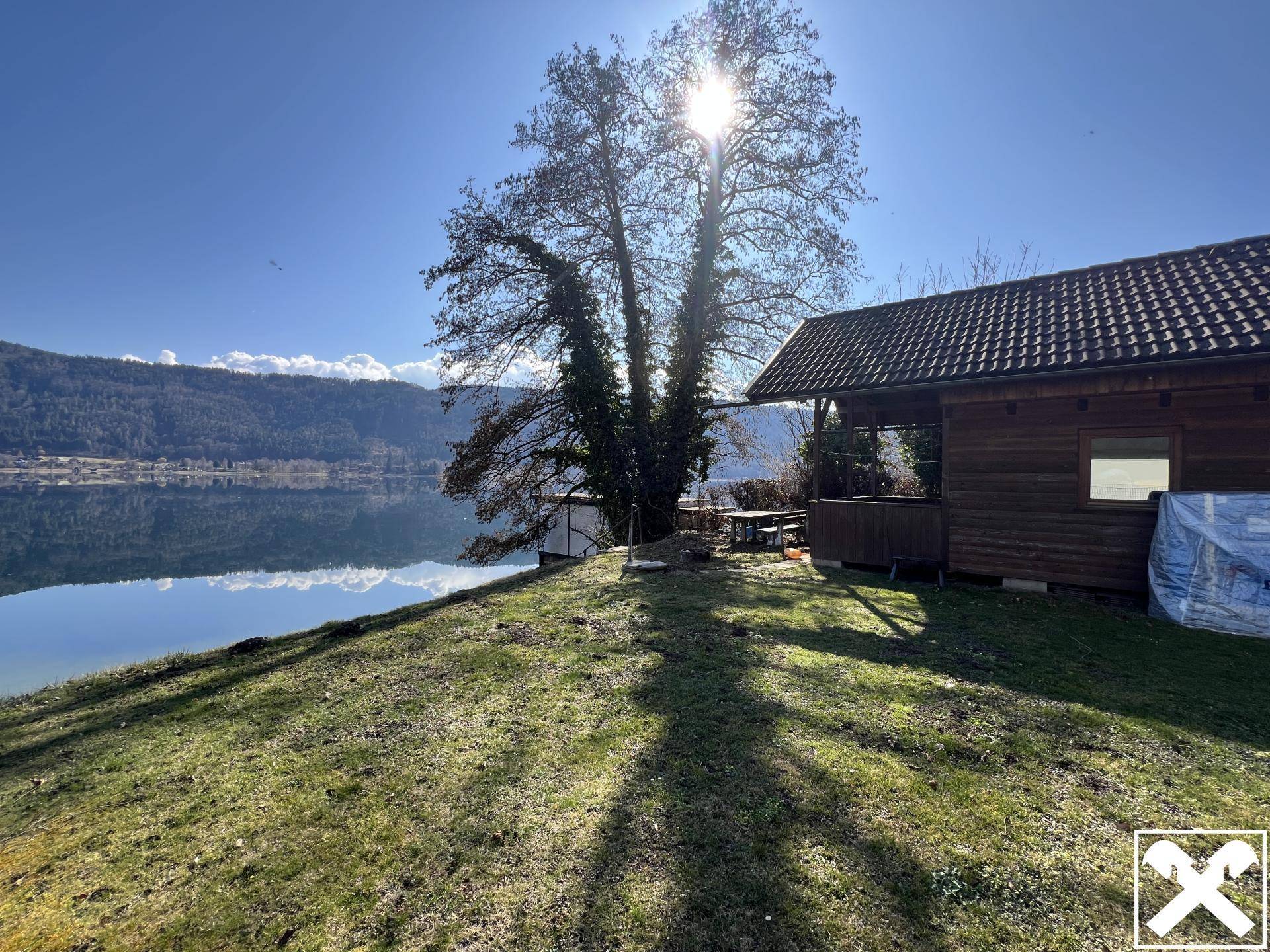 Sonniger Gartenbereich mit altem Baum und kleinem Holzhaus direkt am Seeufer.