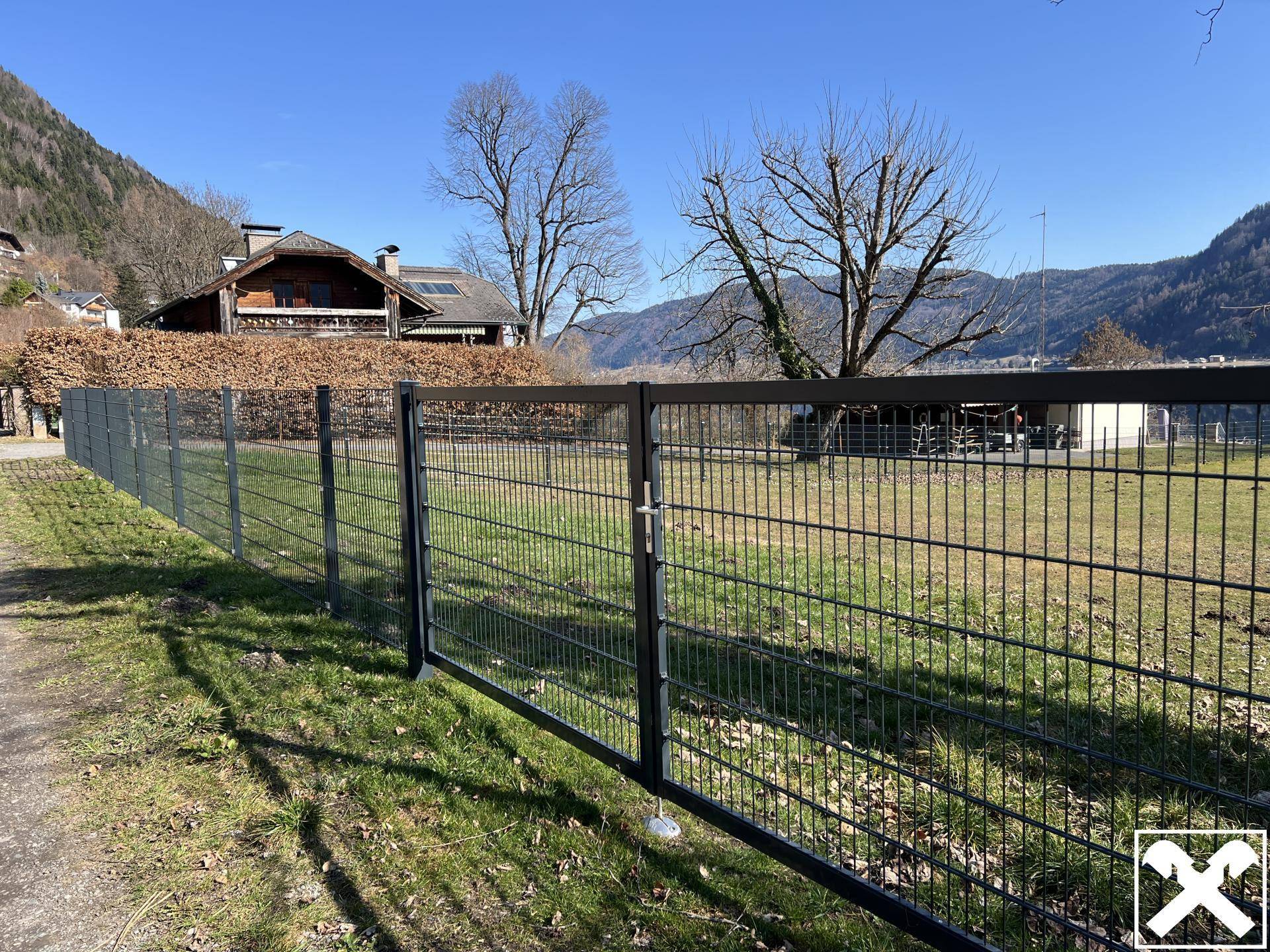 Robuster Metallzaun begrenzt die grüne Rasenfläche mit Blick auf die Häuser und Berge.