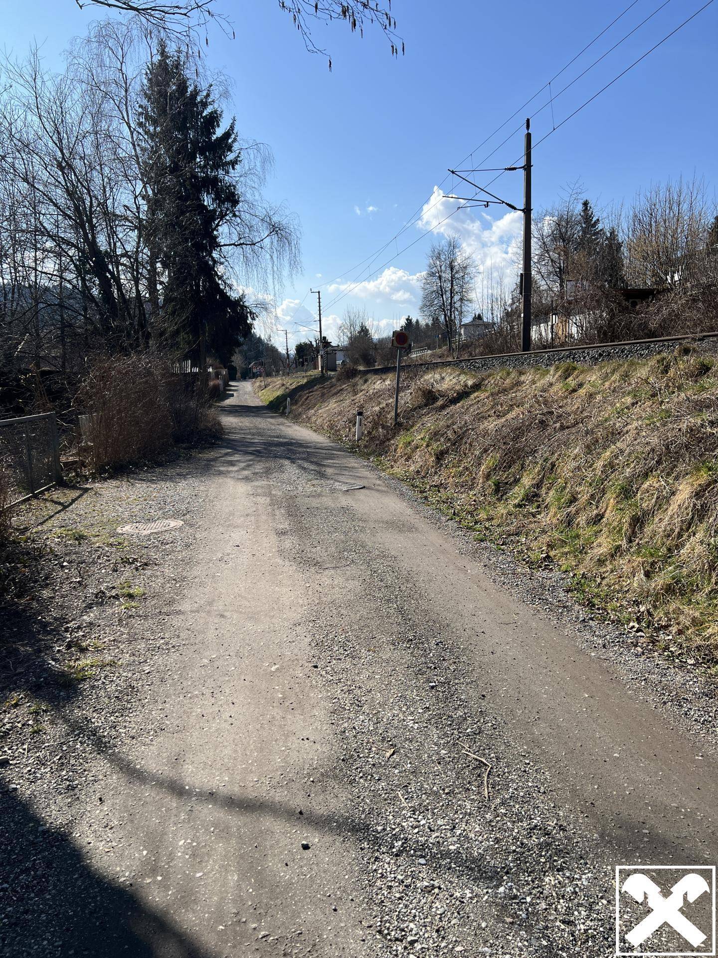 Unbefestigter Weg führt entlang einer Böschung mit Stromleitungen und Bäumen.