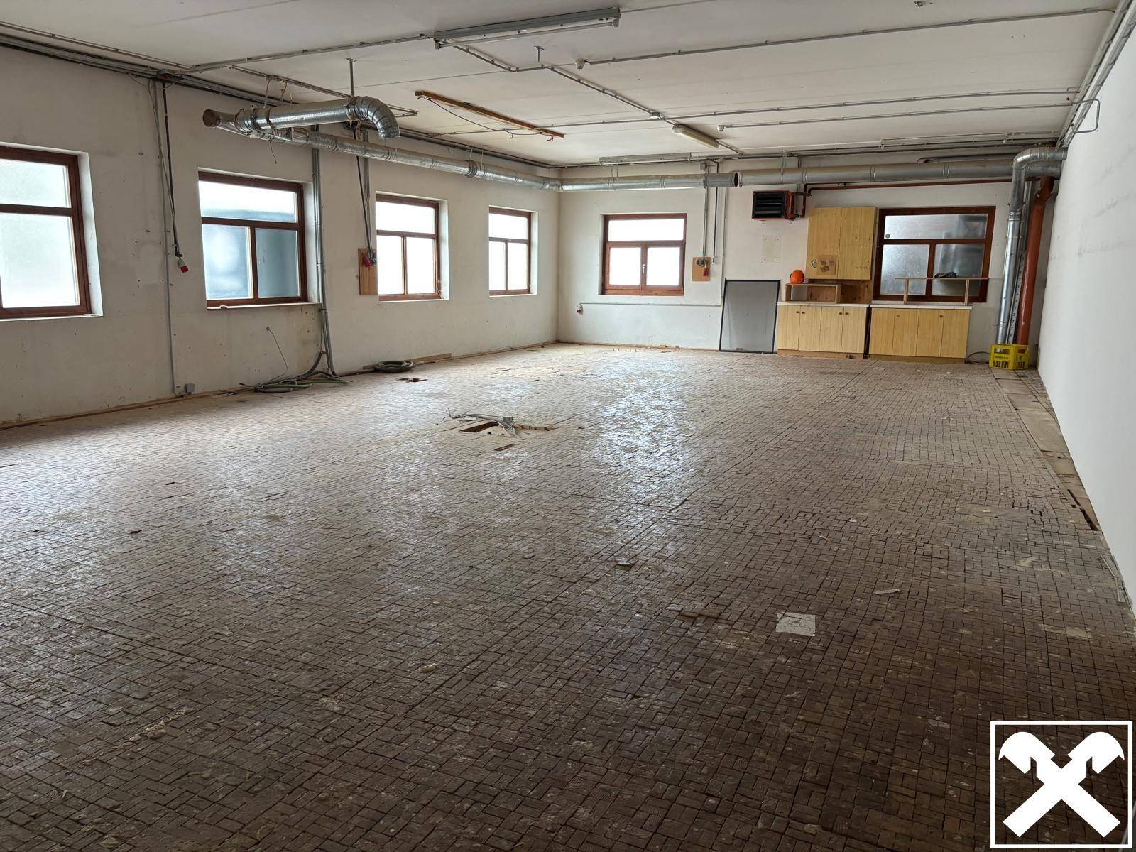 Großer, leerer Raum mit vielen Fenstern, sichtbaren Leitungen und einem Boden aus kleinen Ziegeln oder Fliesen.