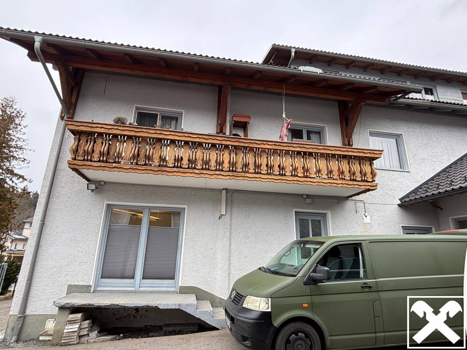 Traditioneller Holzbalkon mit geschnitztem Geländer an einer hell verputzten Fassade, davor ein Lieferwagen.
