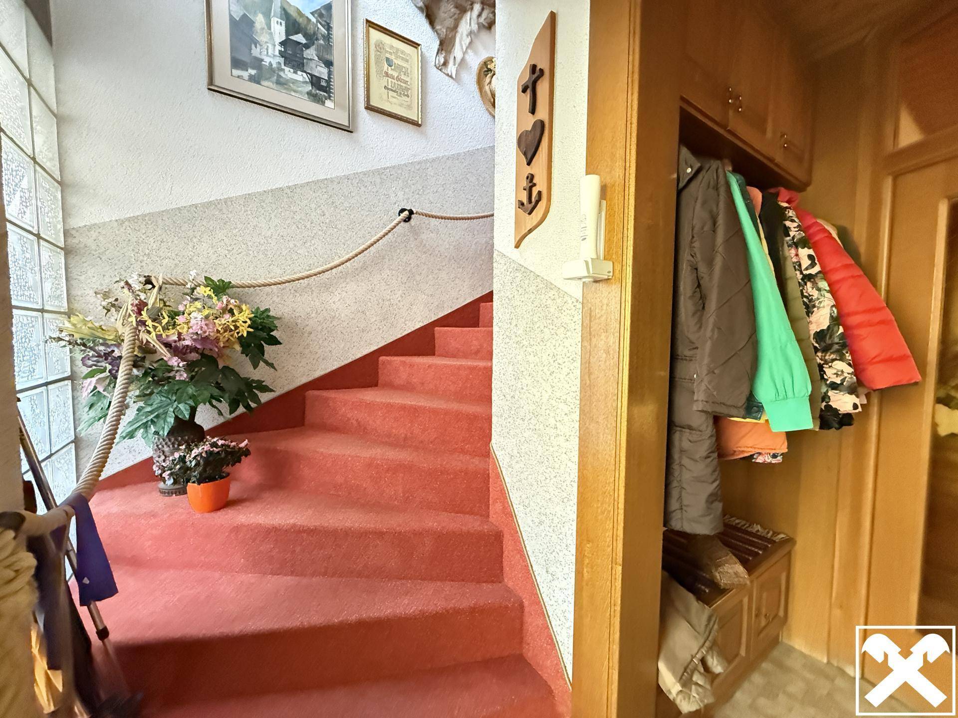 Treppenhaus mit rotem Teppichboden, Holzgeländer und einer Garderobe im Eingangsbereich.
