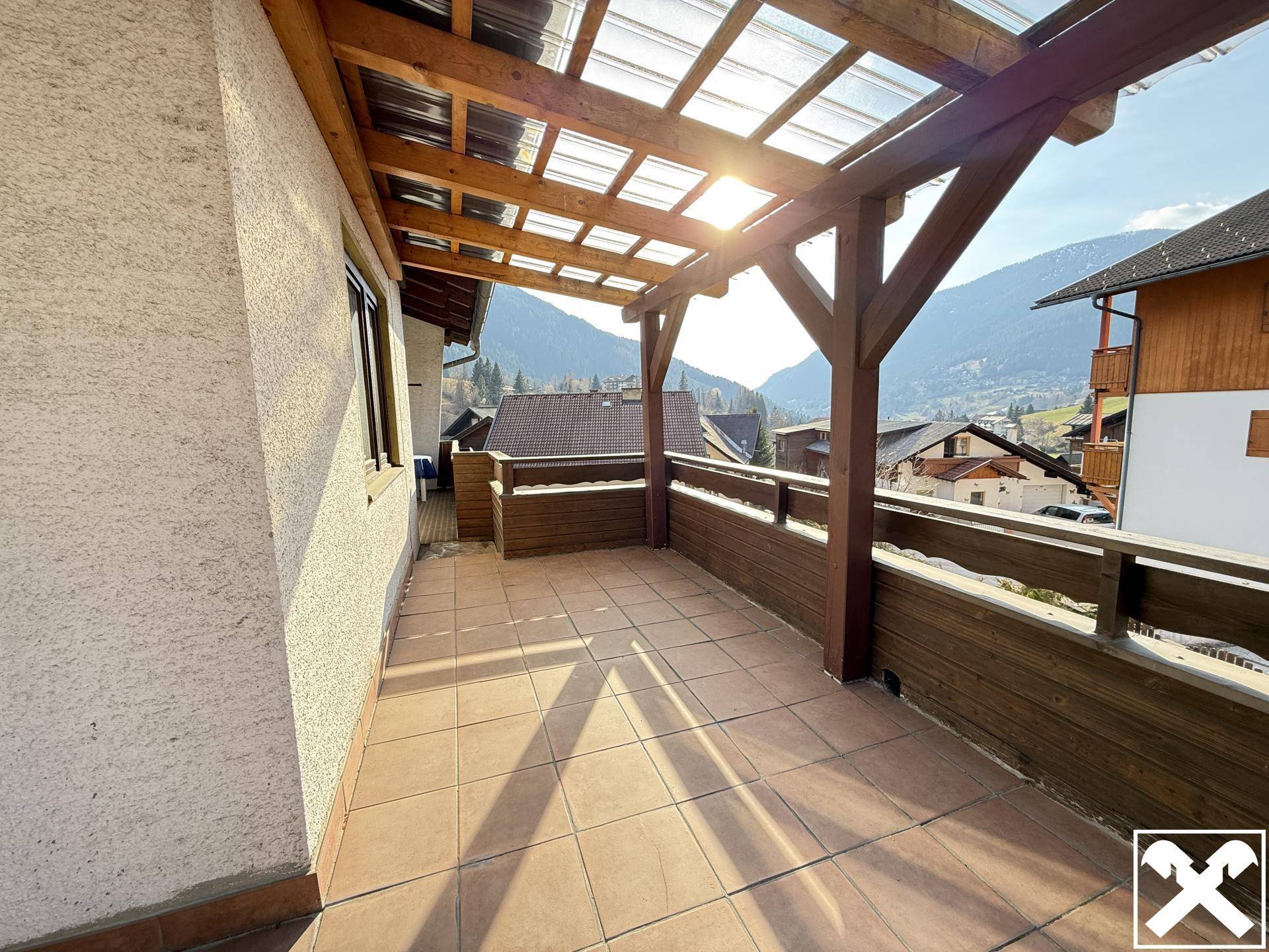 Überdachter Balkon mit Fliesenboden, Holzgeländer und sonnigem Blick auf die Berge.