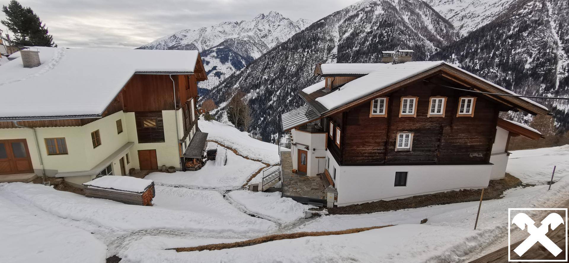 Zwei traditionelle Chalets in einer verschneiten Berglandschaft, umgeben von weiteren Häusern und Natur.