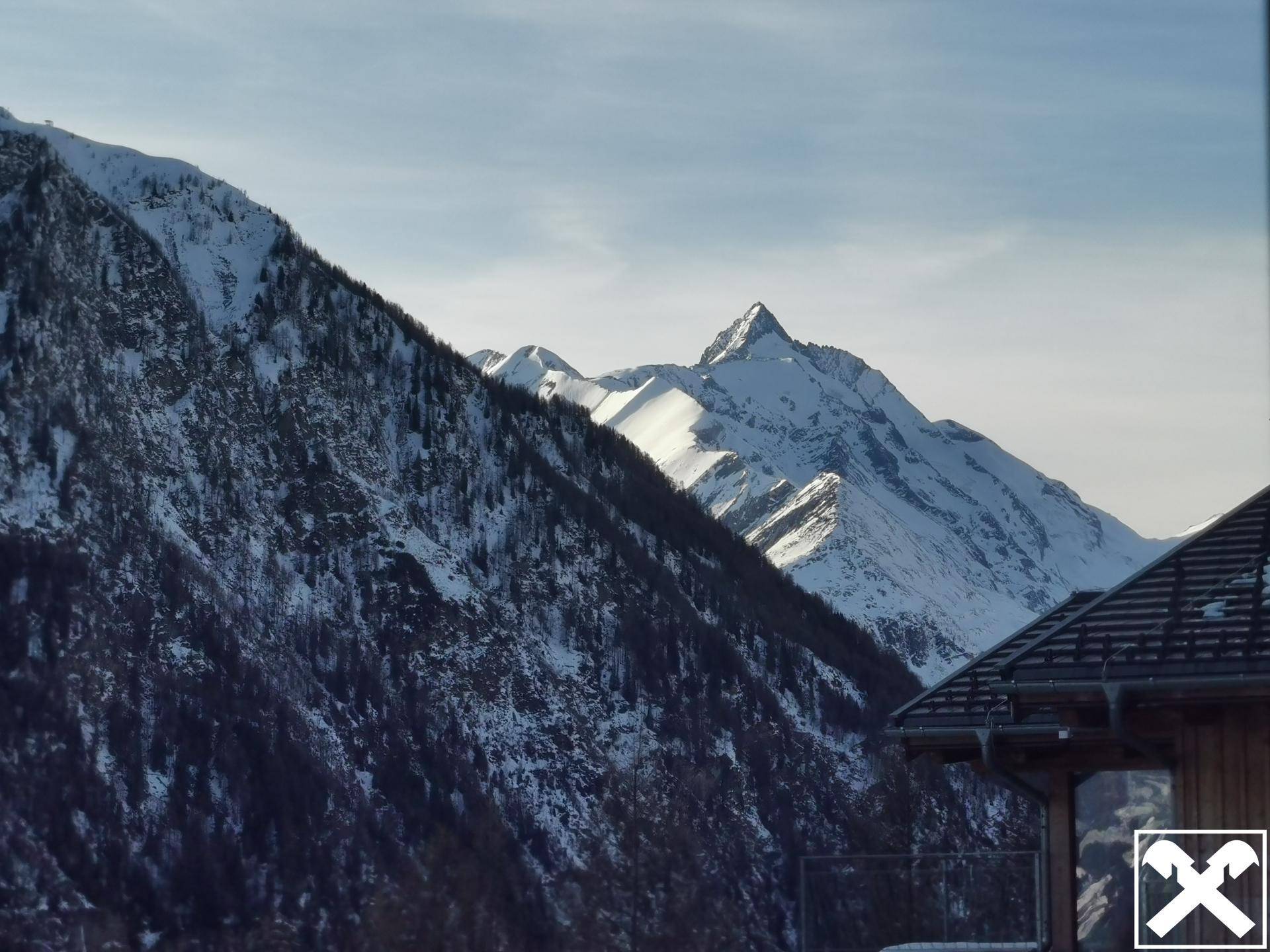 Panoramablick auf schneebedeckte Berge, teilweise verdeckt von einem Gebäudedach im Vordergrund.