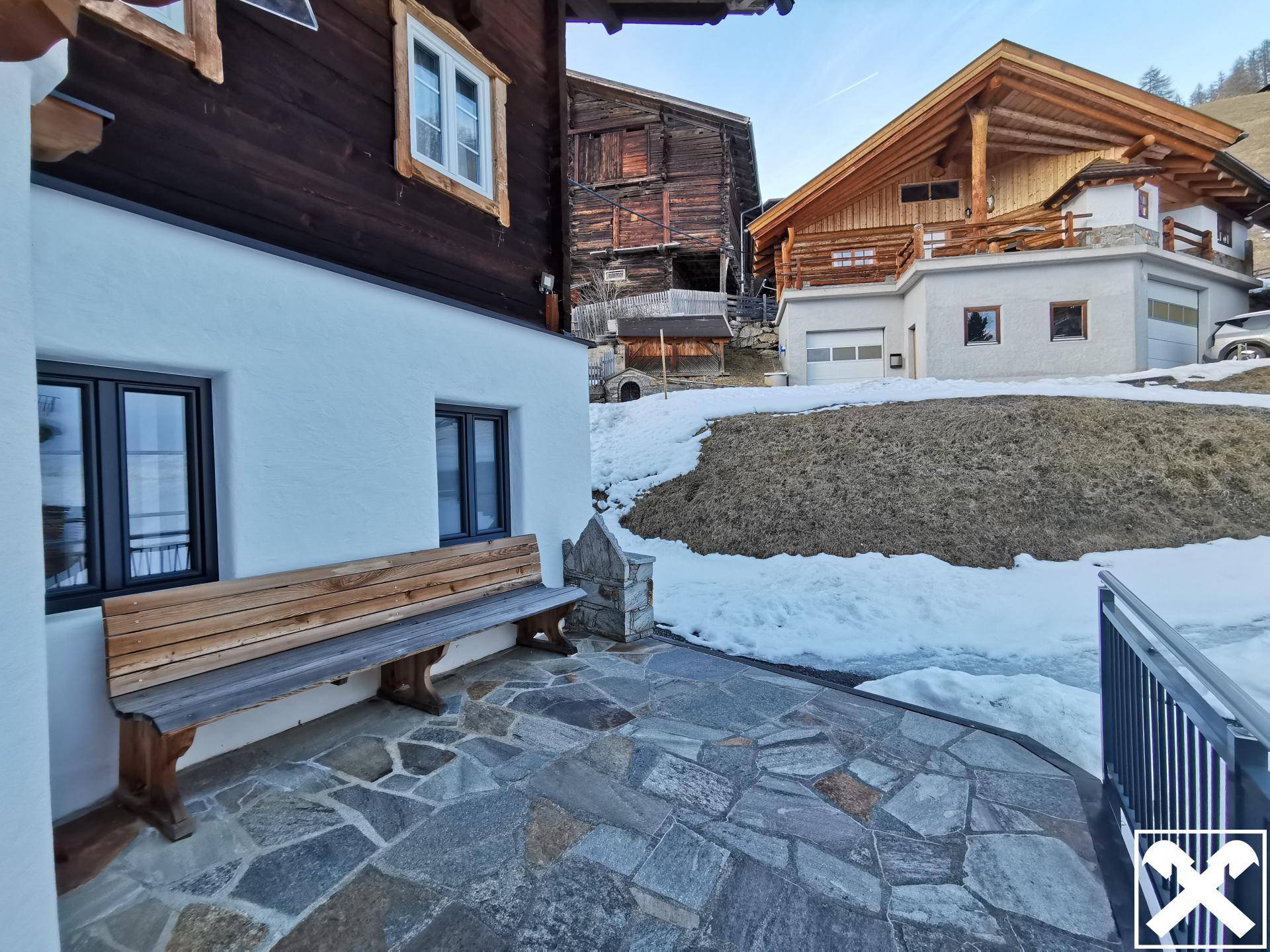 Eingangsbereich mit Steinterrasse und rustikaler Holzbank, umgeben von Schnee und weiteren Gebäuden.