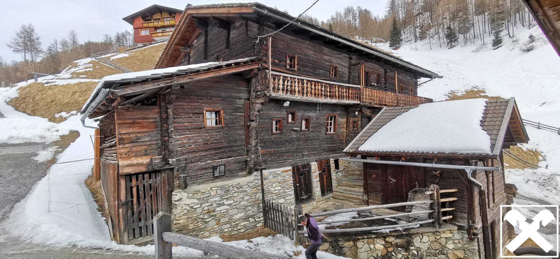Rustikales Holzhaus mit Balkon und Steinsockel in winterlicher Umgebung, umgeben von Schnee.