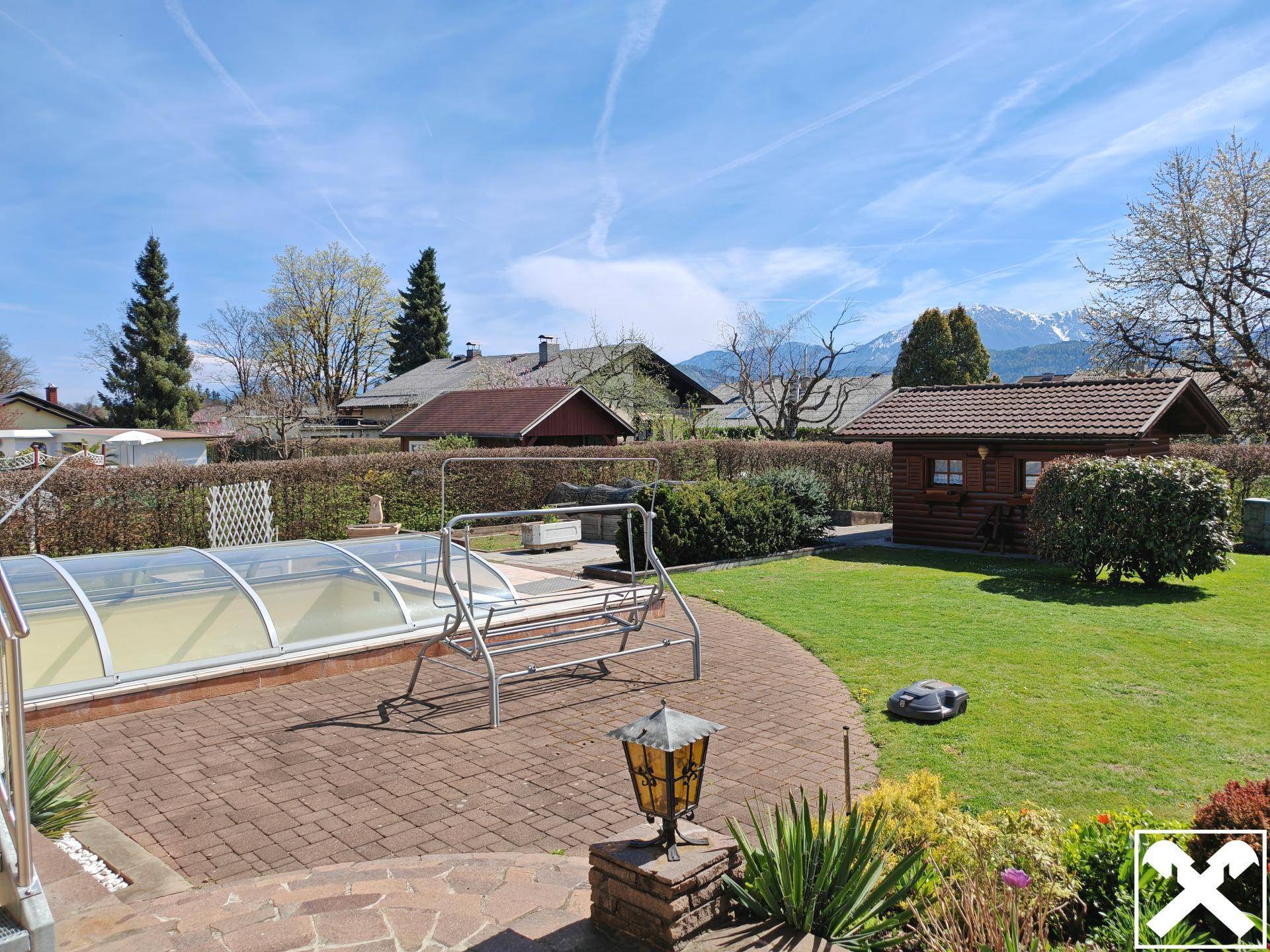 Gartenbereich mit überdachtem Pool, Schaukel und Gartenhaus vor Bergkulisse.