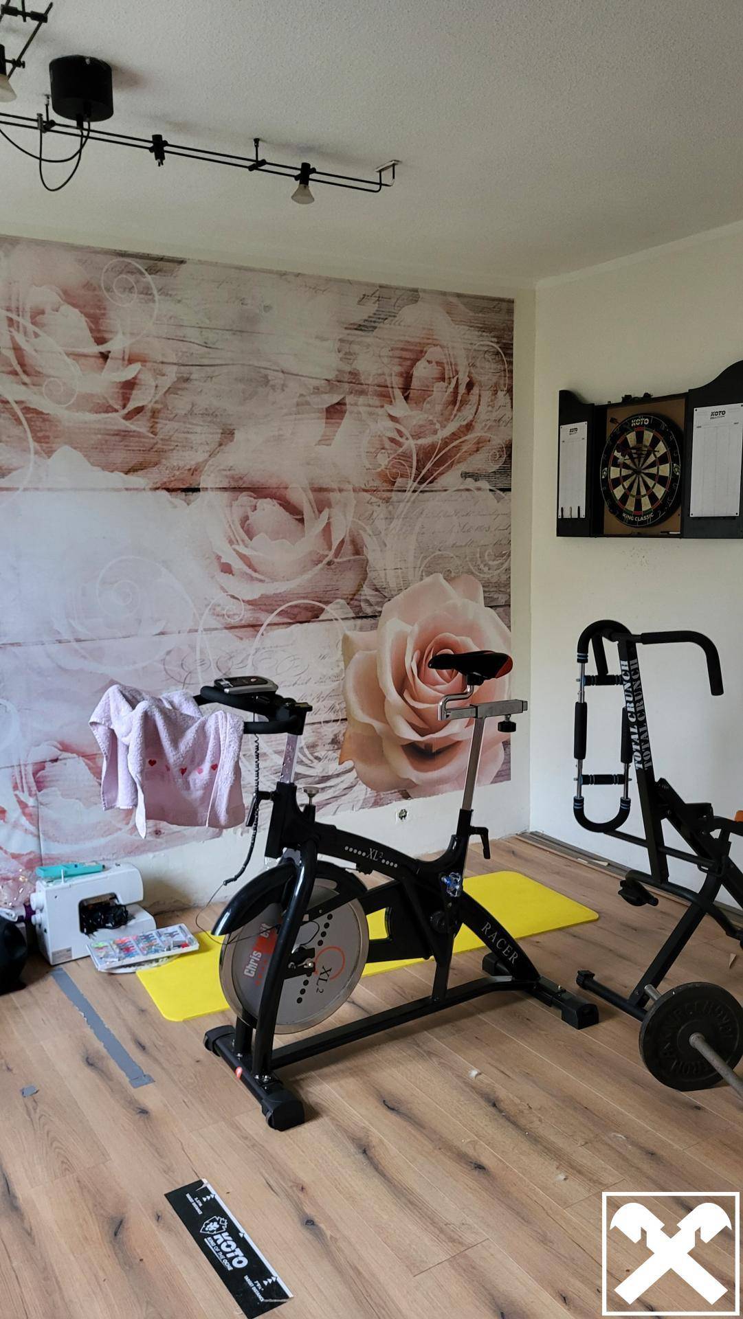 Fitnessraum mit Trainingsgeräten, Holzboden und floraler Wandtapete.