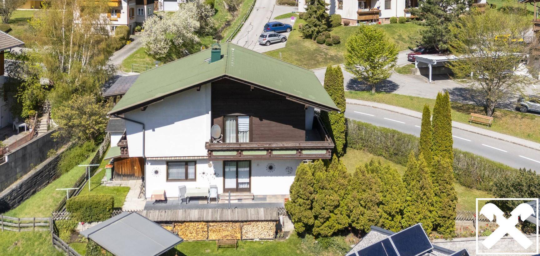 Vogelperspektive eines Hauses mit grünem Dach, umgeben von Bäumen und einer bergigen Landschaft.