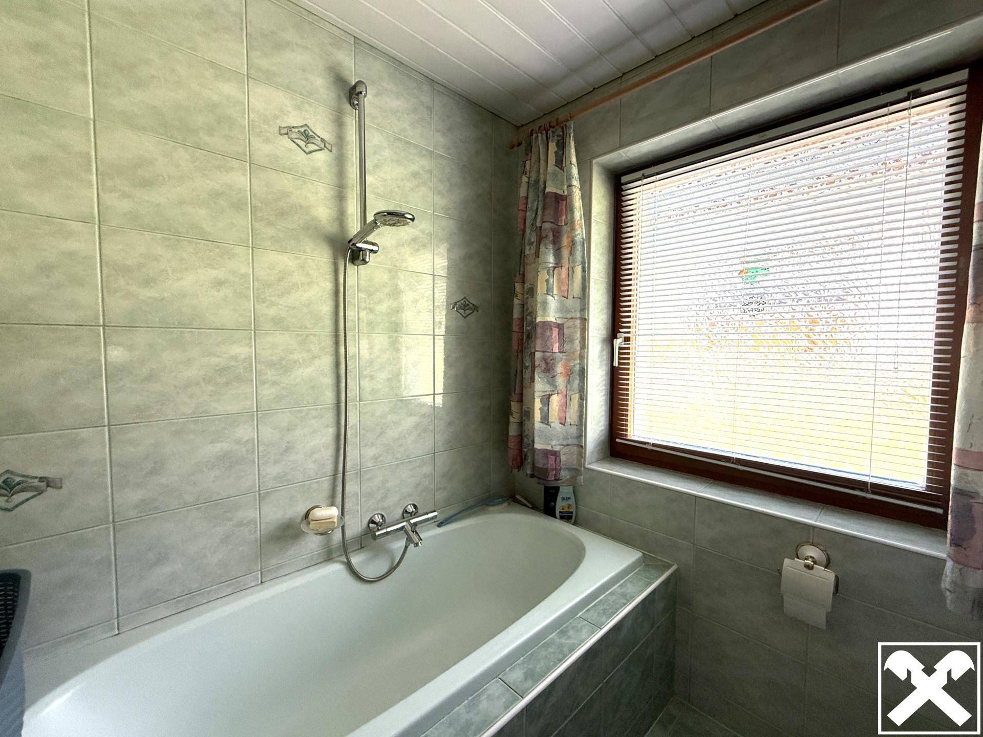 Badezimmer mit Badewanne, Duscharmatur und Fenster, gefliest in hellgrünen Tönen.
