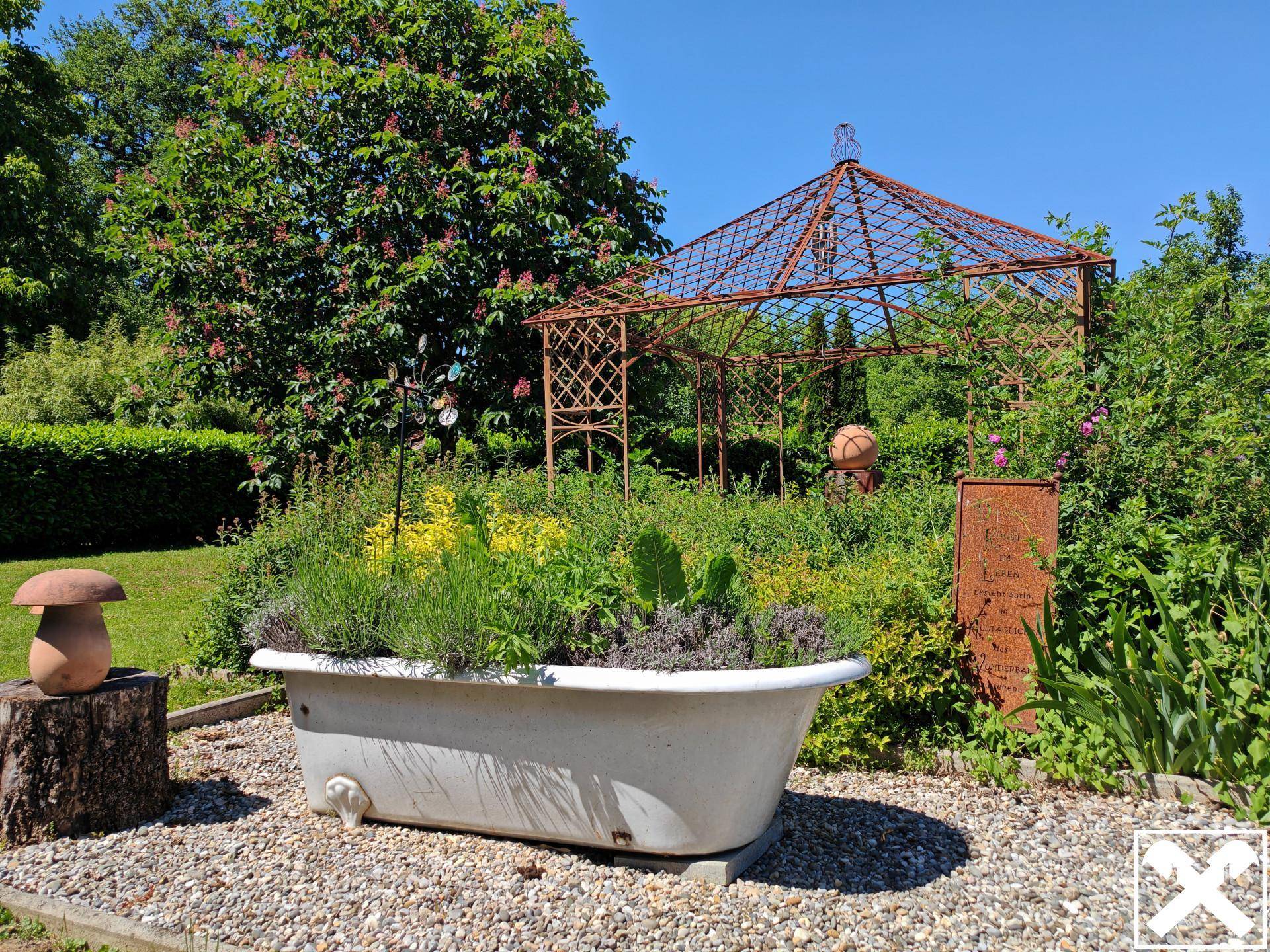 Kreativ gestalteter Gartenbereich mit einer bepflanzten Badewanne und einem Metallpavillon.