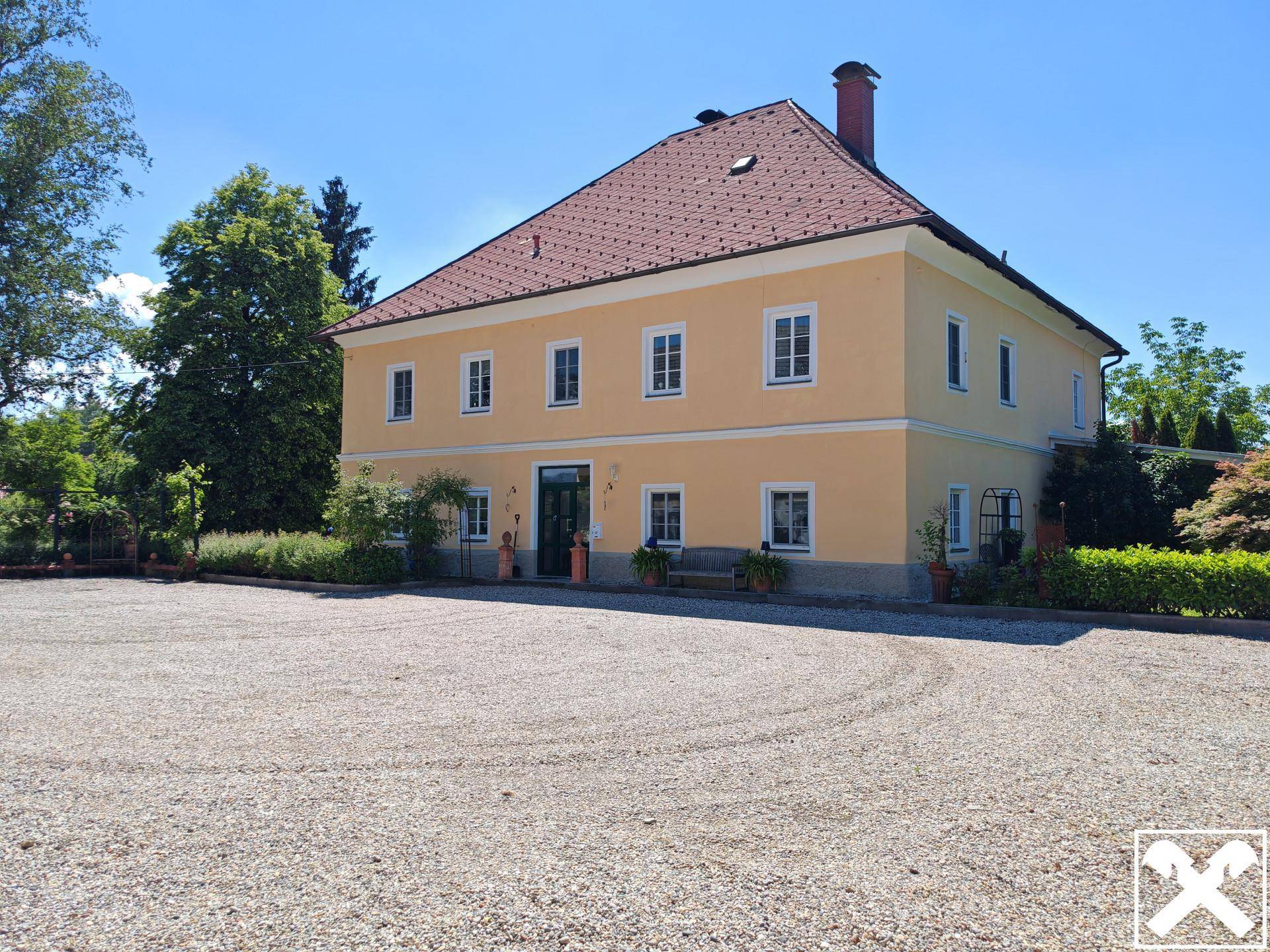 Historisches gelbes Gebäude mit rotem Dach und großem Schottervorplatz unter strahlend blauem Himmel.