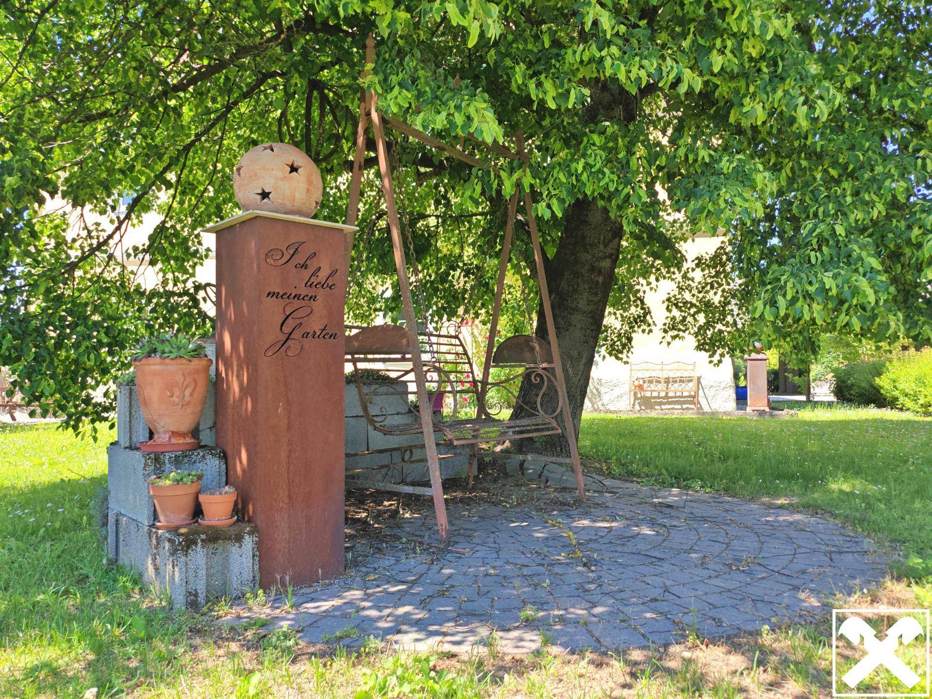 Gemütlicher Sitzbereich mit Schaukel unter einem Baum und dekorativem Pfeiler im Garten.